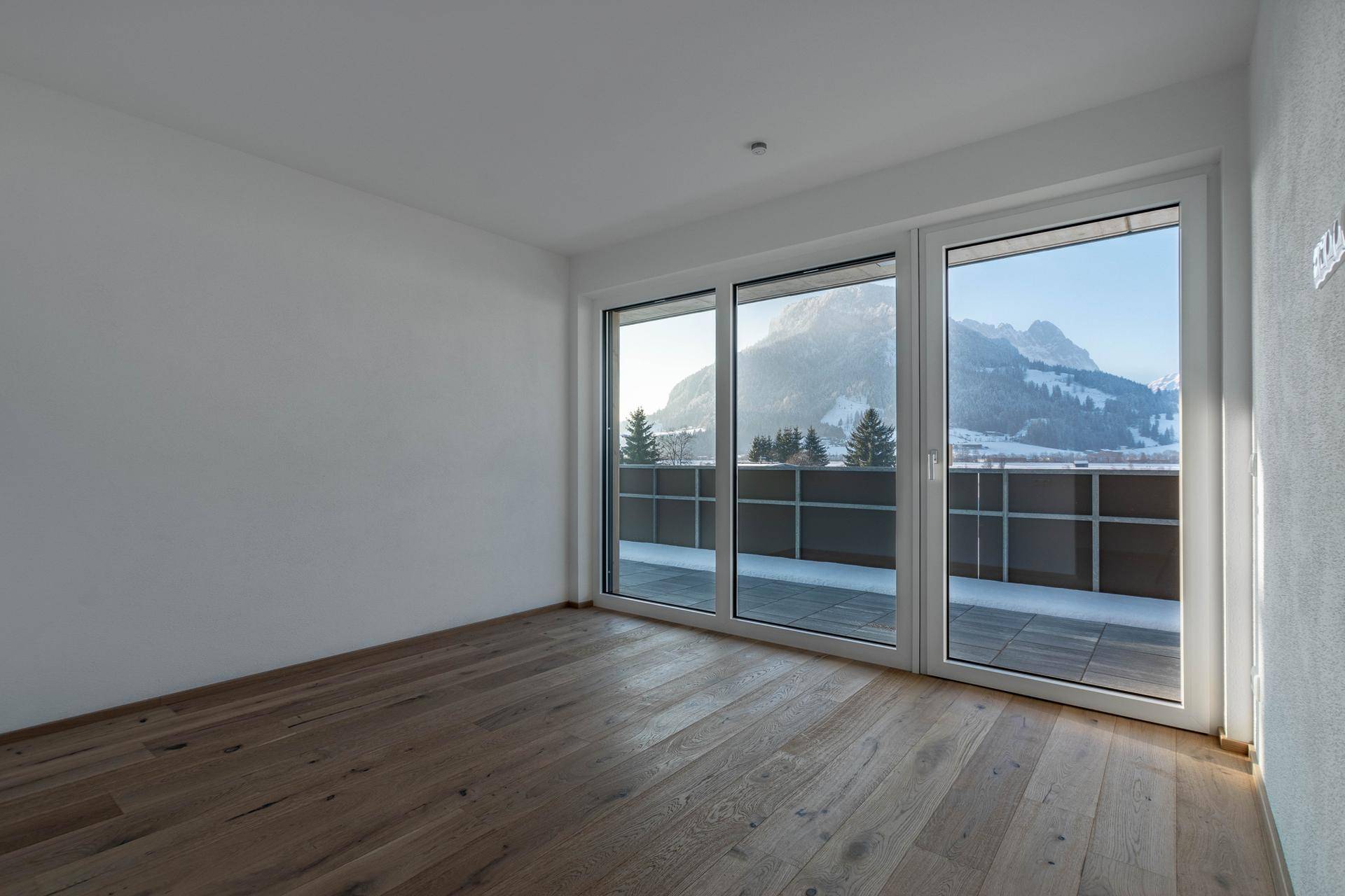 Heller Raum mit Holzboden und großen Fenstern, die einen weiten Blick auf die verschneiten Berge bieten.