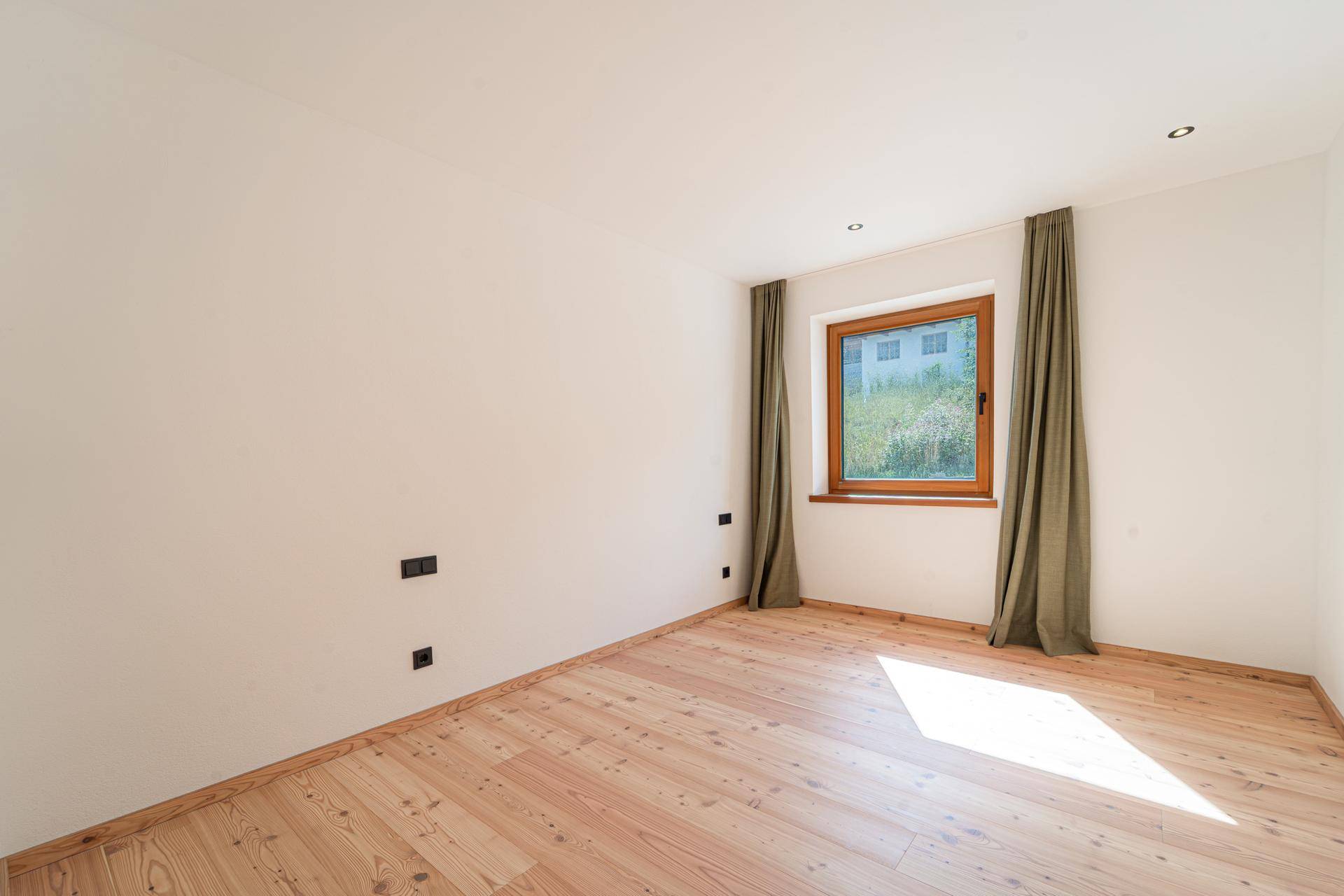 Helles Schlafzimmer mit Holzboden und großem Fenster, das viel Tageslicht hereinlässt.