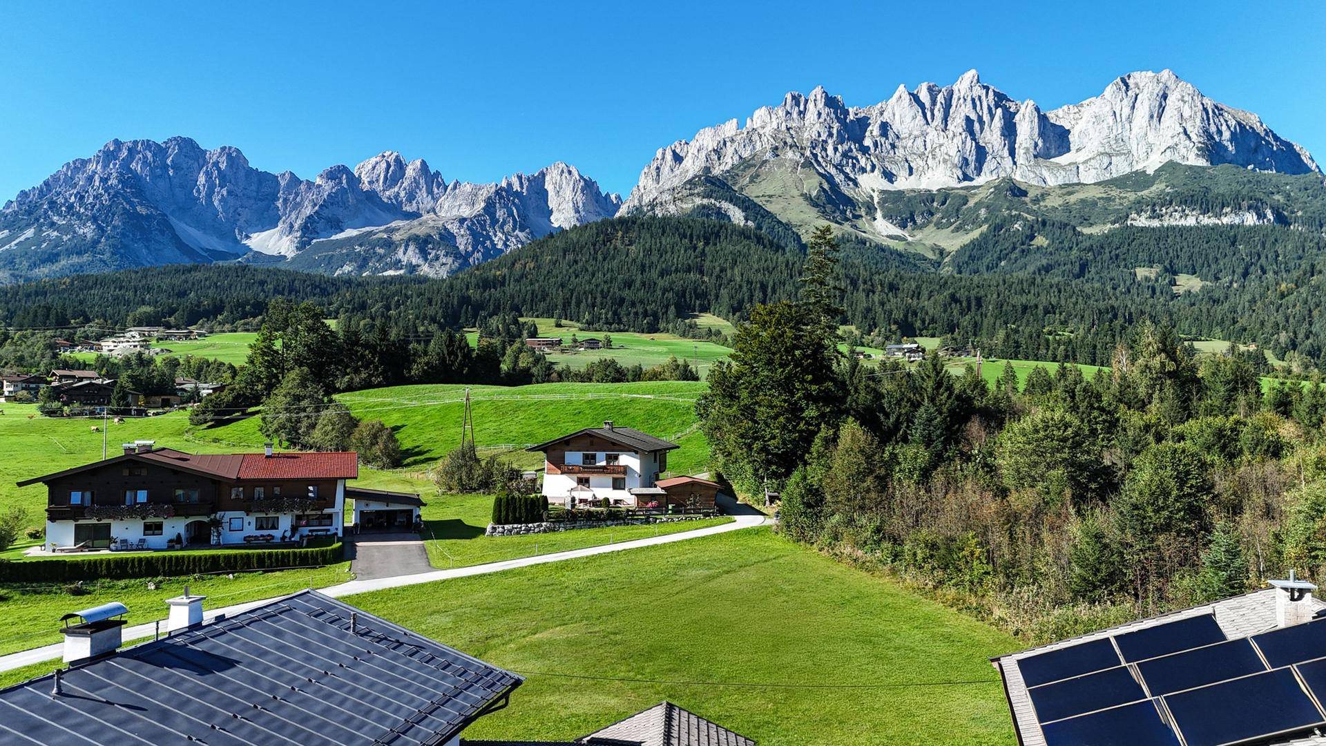 Weitläufiger Blick über grüne Wiesen und Häuser mit beeindruckender Bergkette im Hintergrund unter blauem Himmel.