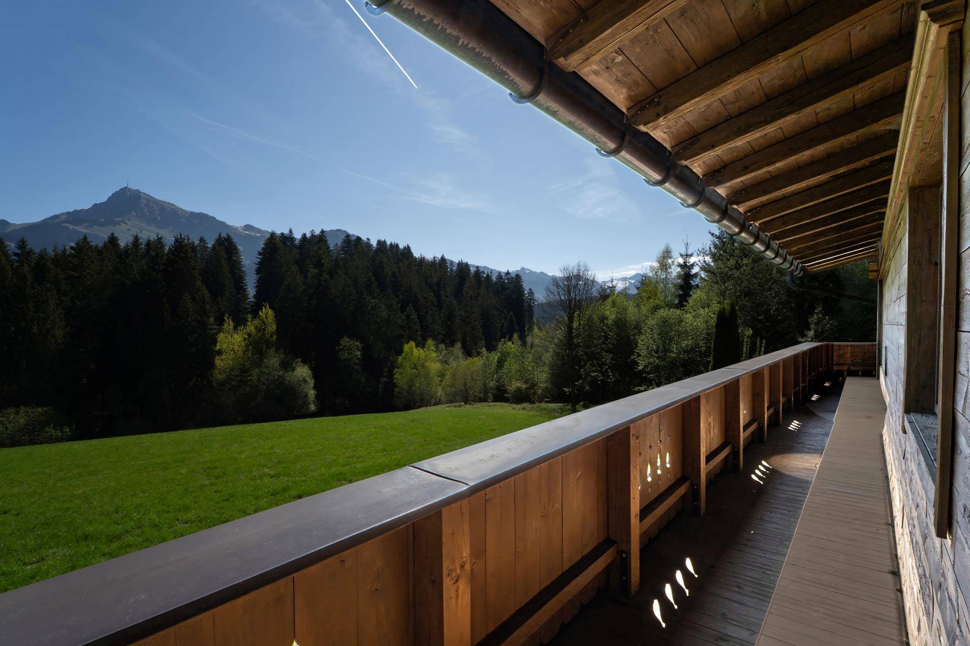 Langer Holzbalkon mit Blick auf grüne Wiesen und bewaldete Berge unter blauem Himmel.