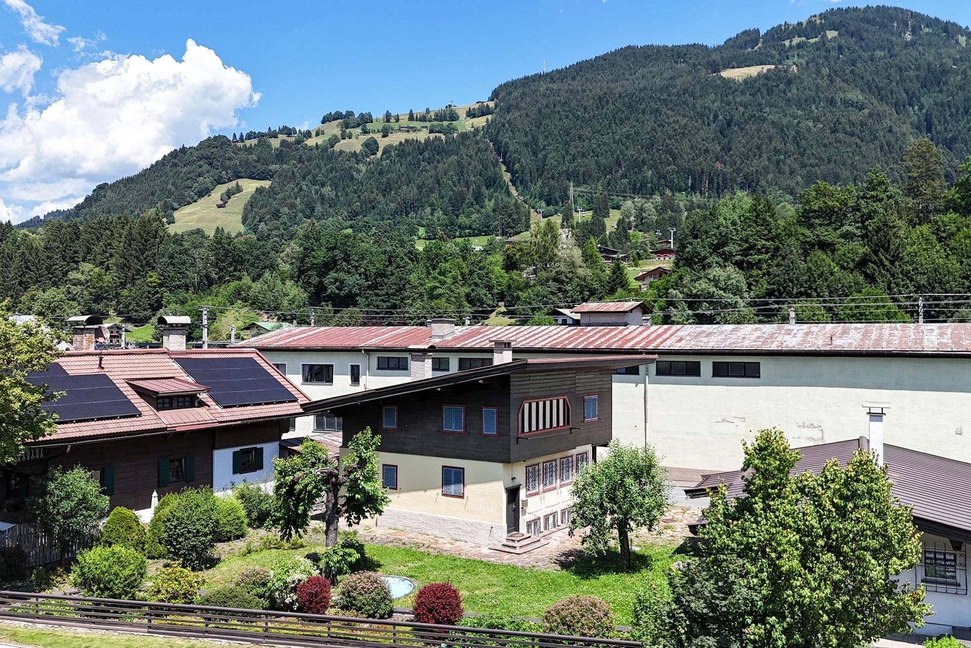 Gebäude mit Solaranlagen auf dem Dach, umgeben von grünen Hügeln und weiteren Häusern.