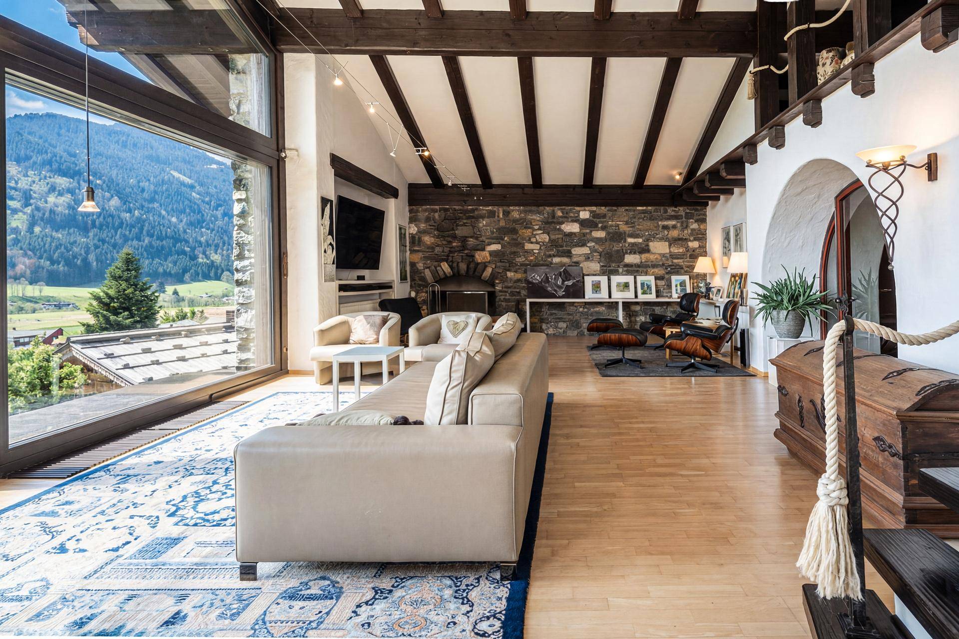 Geräumiges Wohnzimmer mit Steinwand, Holzbalken und großen Fenstern mit Blick auf die Berge.