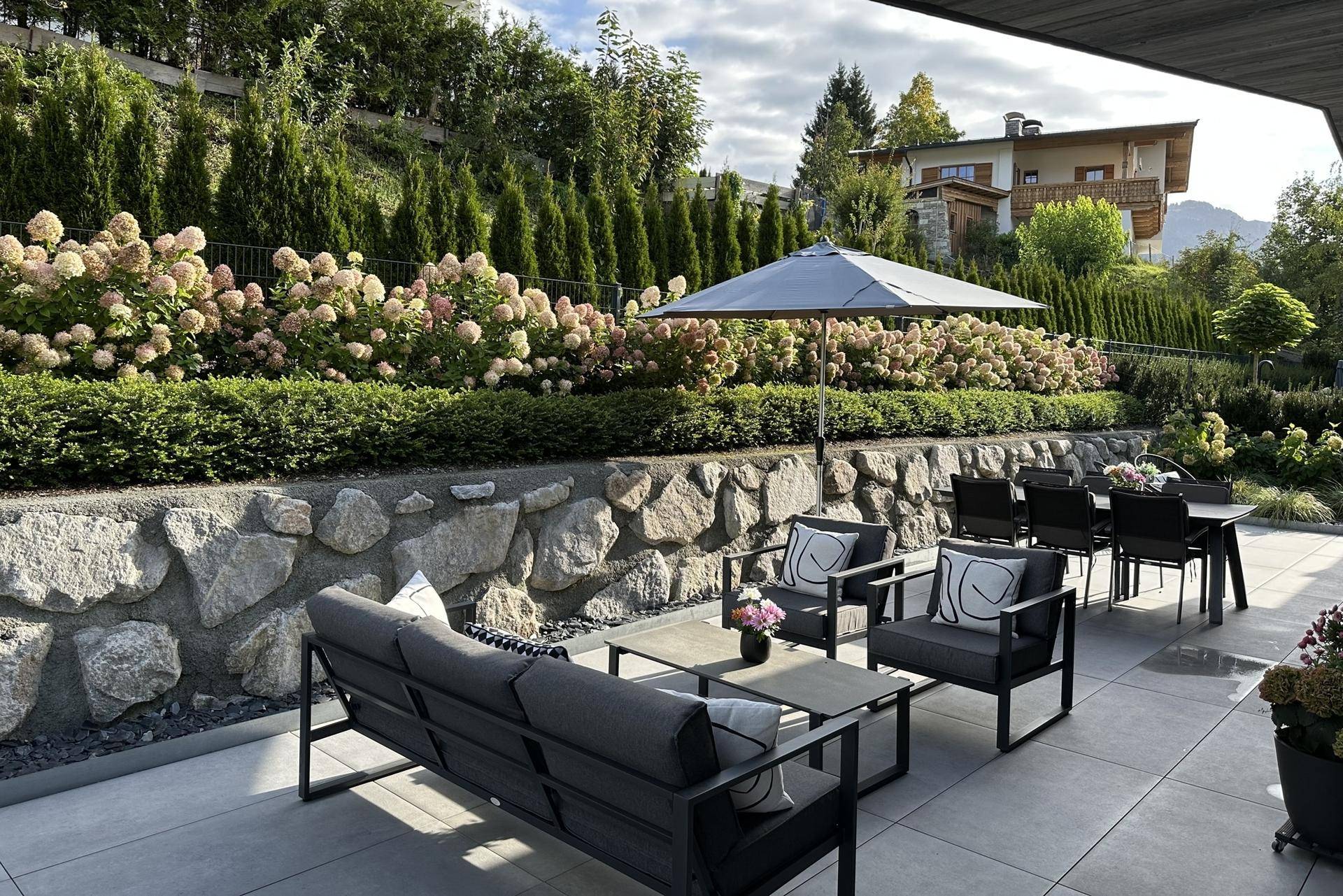 Großzügige Terrasse im Erdgeschoss mit Lounge-Möbeln, Sonnenschirm und einer bepflanzten Steinmauer.