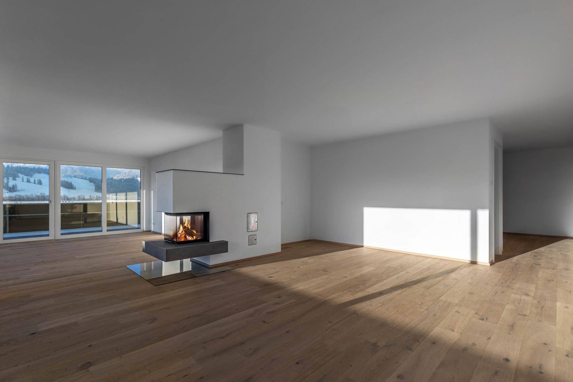 Heller, leerer Wohnbereich mit modernem Kamin, Holzboden und großen Fenstern mit Bergblick.