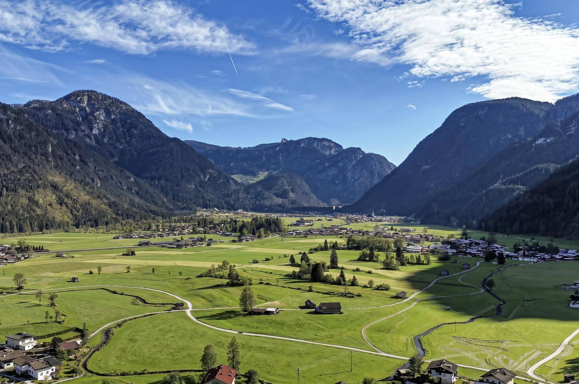 Weitläufige Luftaufnahme eines grünen Tals mit einem Dorf, Feldern und majestätischen Bergen unter klarem Himmel.