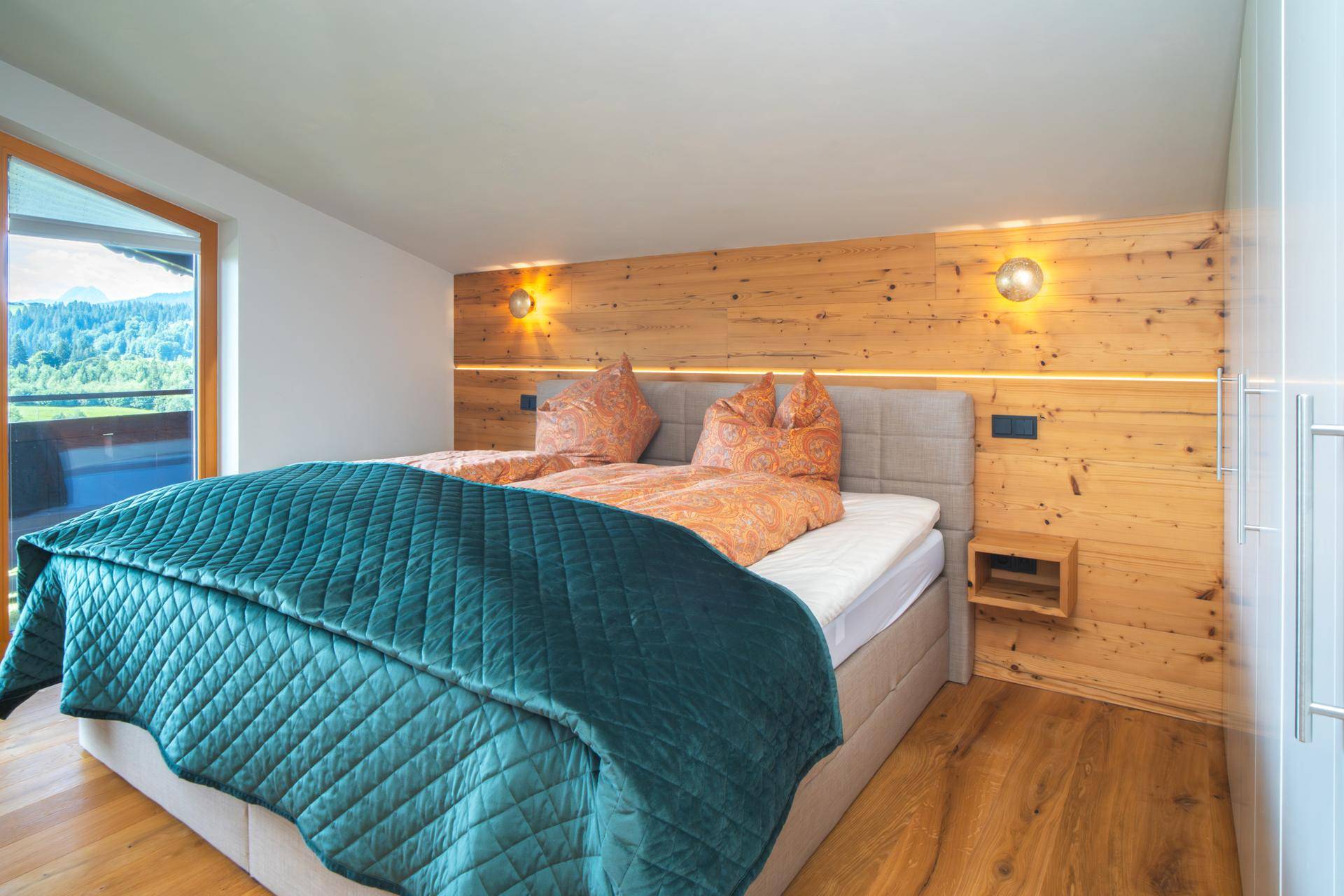 Helles Schlafzimmer mit Holzwand, Doppelbett und direktem Zugang zum Balkon mit Bergblick.