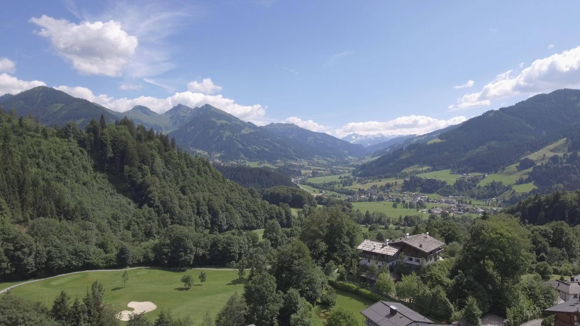 Panoramablick über ein weites grünes Tal mit Golfplatz und Häusern, umgeben von majestätischen Bergen.