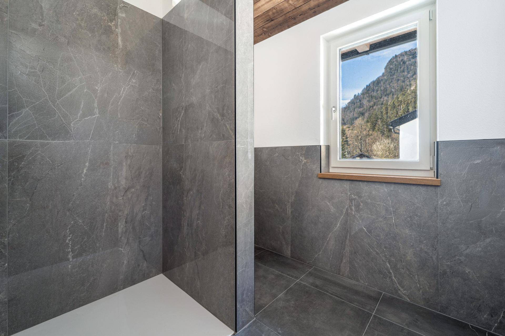 Modernes Badezimmer mit begehbarer Dusche, grauen Fliesen und einem Fenster mit Bergblick.