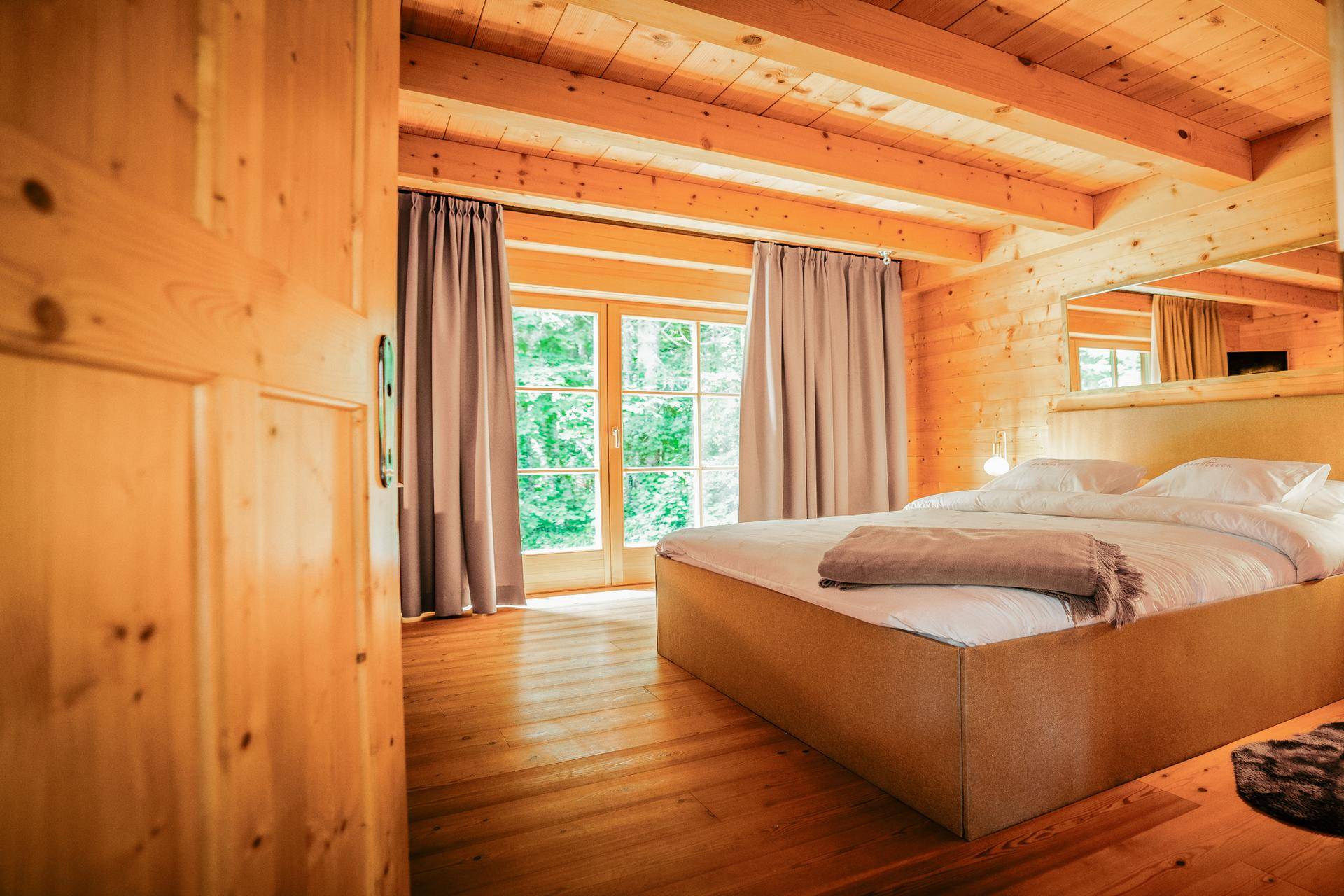 Helles Schlafzimmer mit Holzboden, Holzwänden und großen Fenstern mit Blick ins Grüne.
