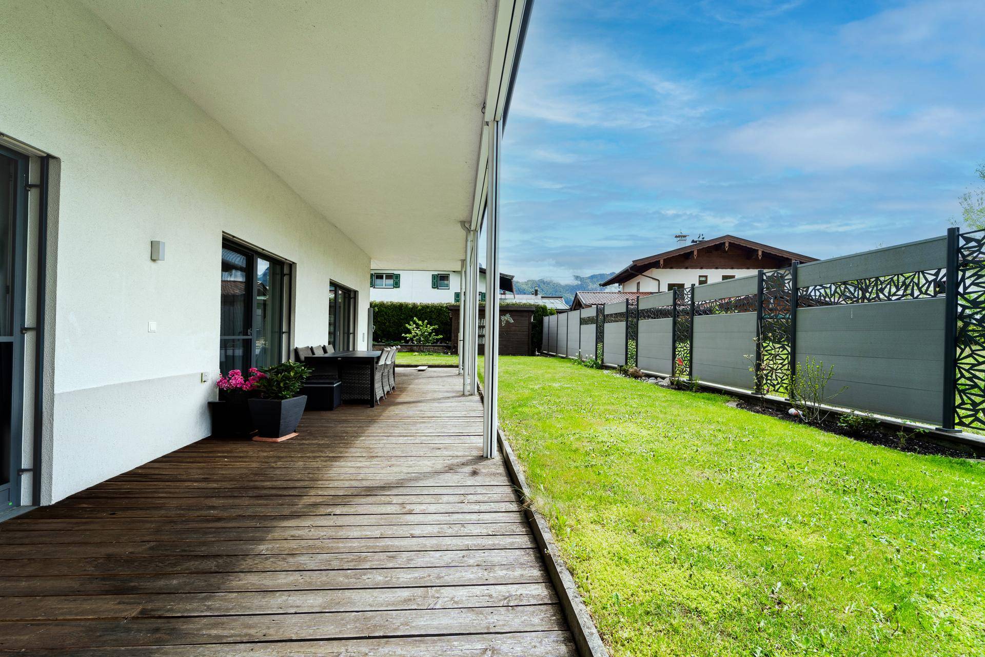 Lange Holzterrasse mit Gartenmöbeln und Blick auf den umzäunten Gartenbereich.