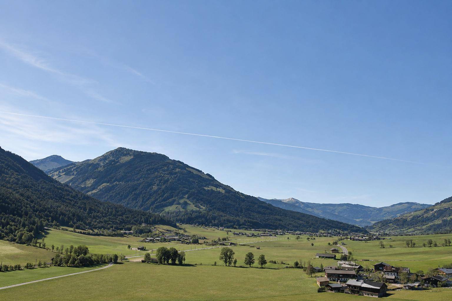 Panoramablick auf die majestätische Bergwelt und weite grüne Wiesen unter blauem Himmel.