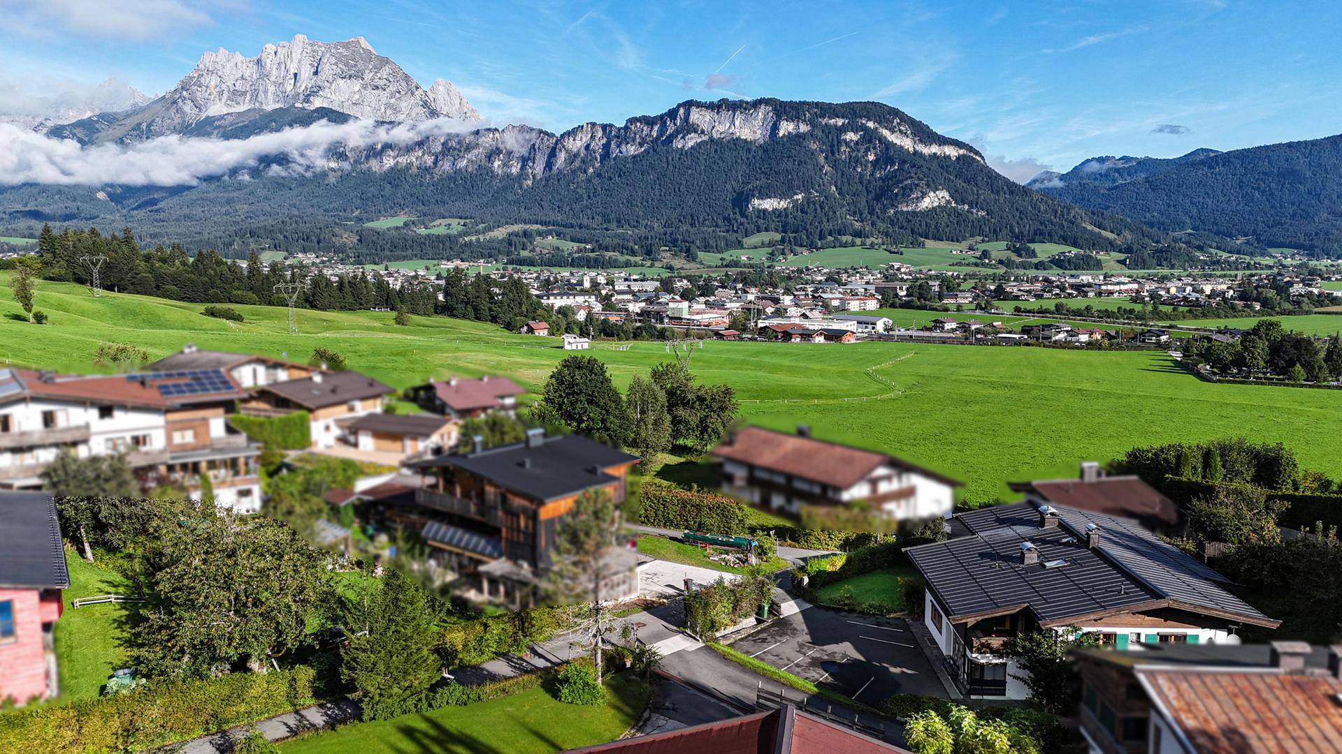 Weitläufige Luftaufnahme der Wohnsiedlung mit grünen Feldern und majestätischen Bergen im Hintergrund.
