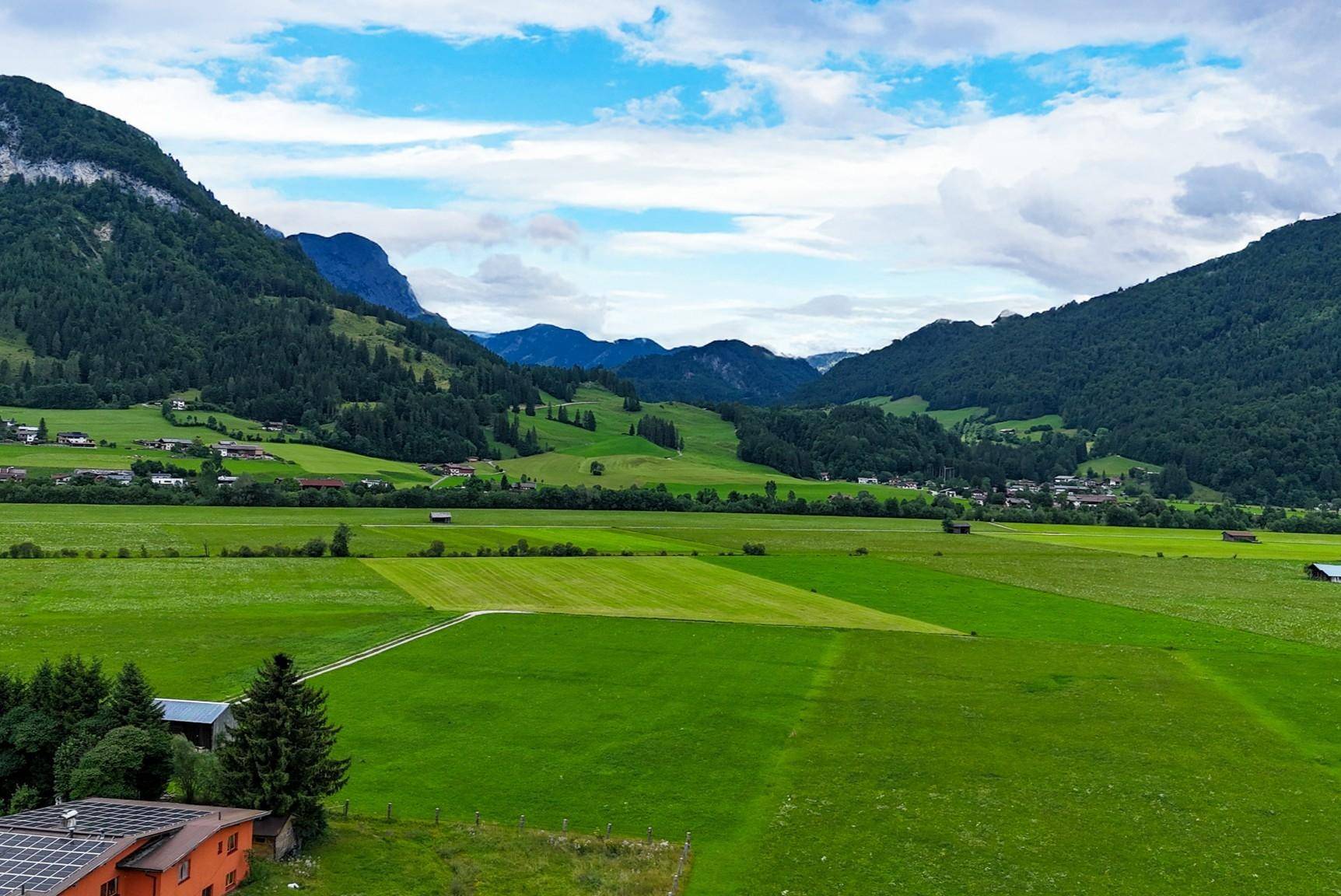 Weitläufige grüne Wiesen und Felder mit bewaldeten Bergen unter einem bewölkten Himmel.