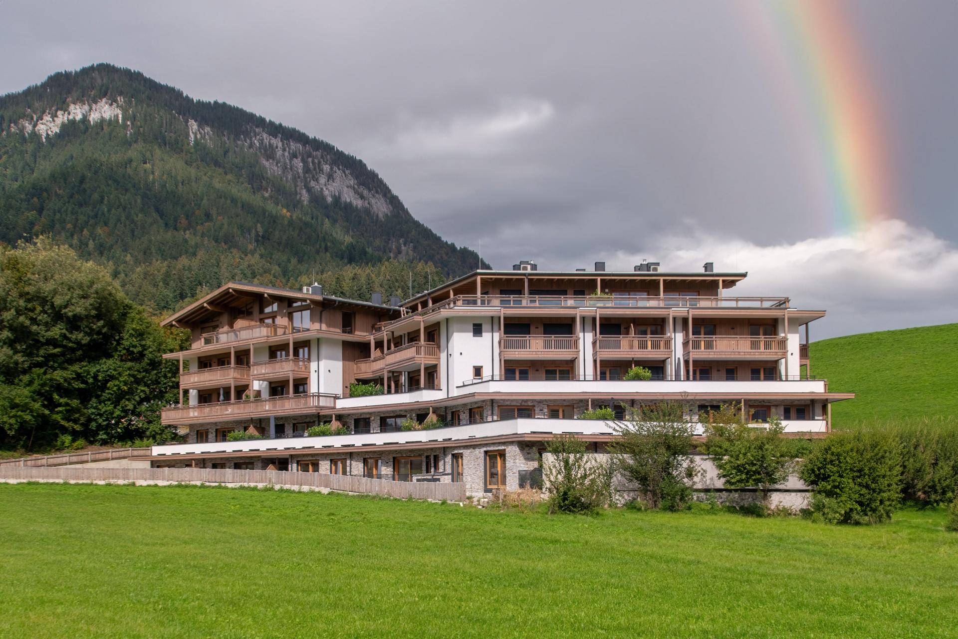 Attraktives Mehrfamilienhaus in alpiner Architektur, eingebettet in eine grüne Landschaft mit Bergblick und Regenbogen.