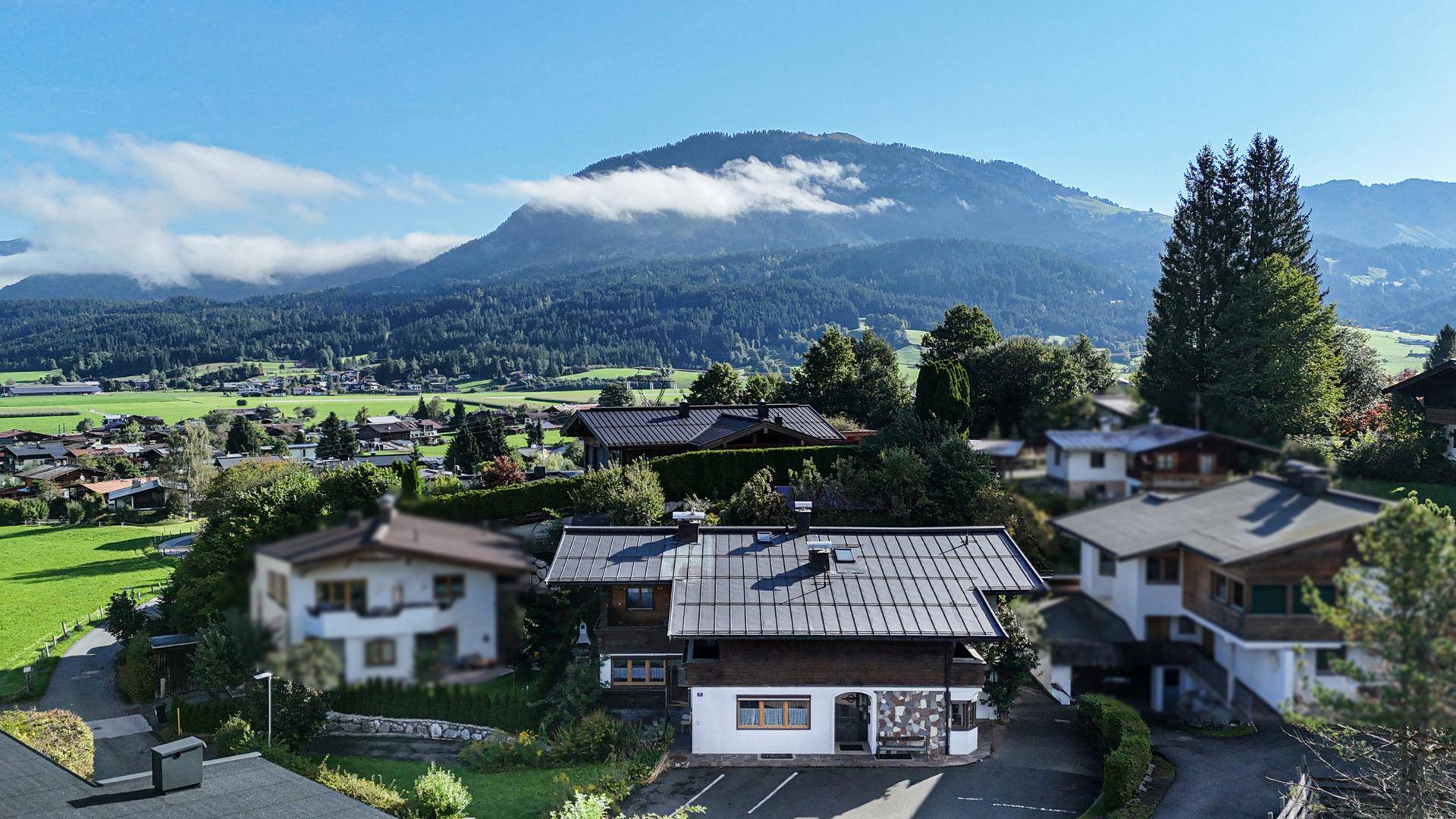 Luftaufnahme eines Hauses mit dunklem Dach und umliegenden Gebäuden, eingebettet in eine grüne Berglandschaft.