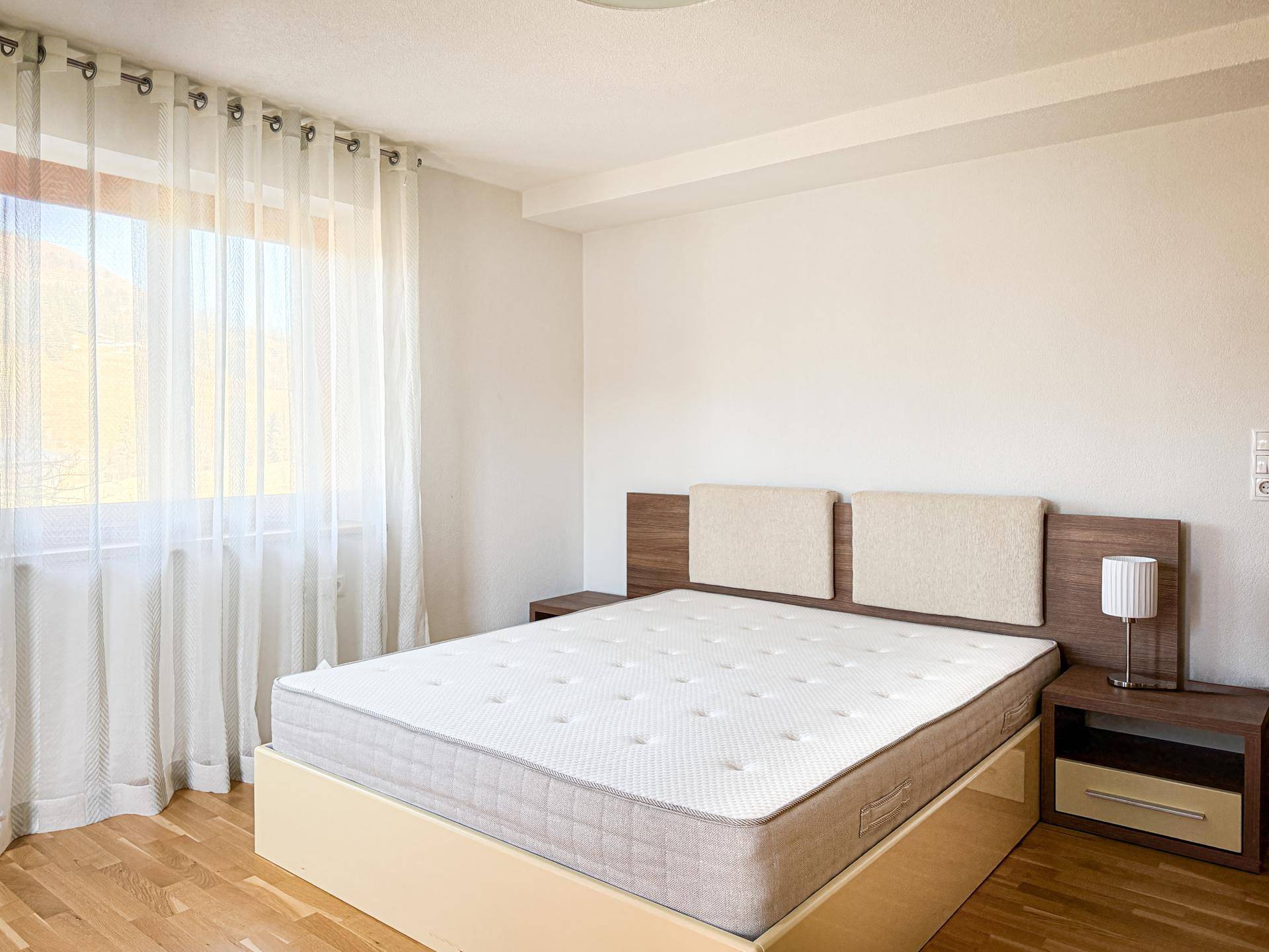 Helles Schlafzimmer mit Holzboden, großem Fenster und schlichtem Doppelbett.
