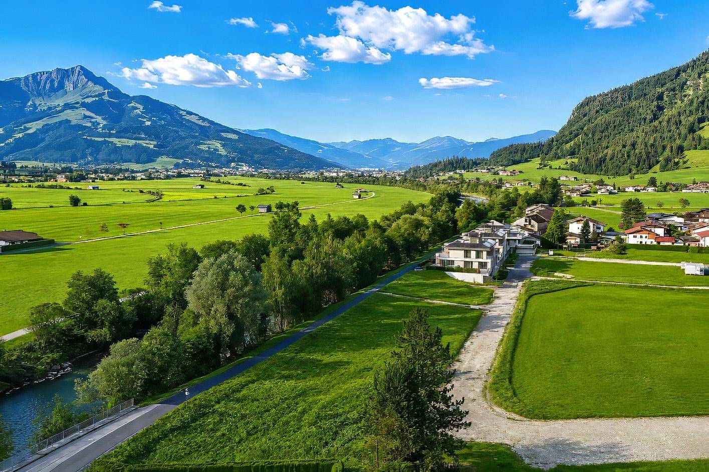 Weitläufige Landschaftsaufnahme mit Fluss, grünen Feldern und modernen Häusern vor Bergkulisse.