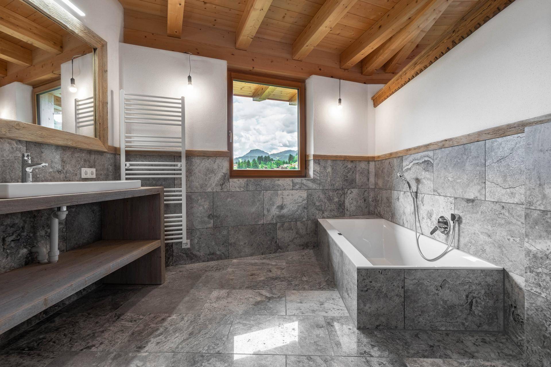 Modernes Badezimmer mit Badewanne, Holzdecke, Fliesenboden und Fenster mit Bergblick.
