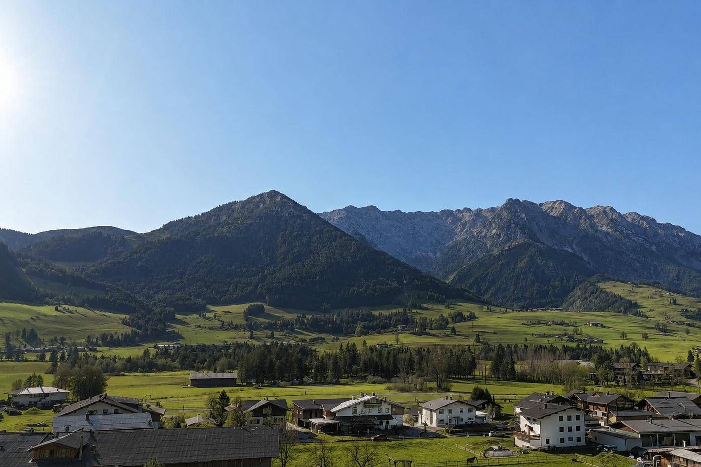Weitläufiger Blick über ein grünes Tal mit verstreuten Häusern und majestätischen bewaldeten Bergen im Hintergrund.