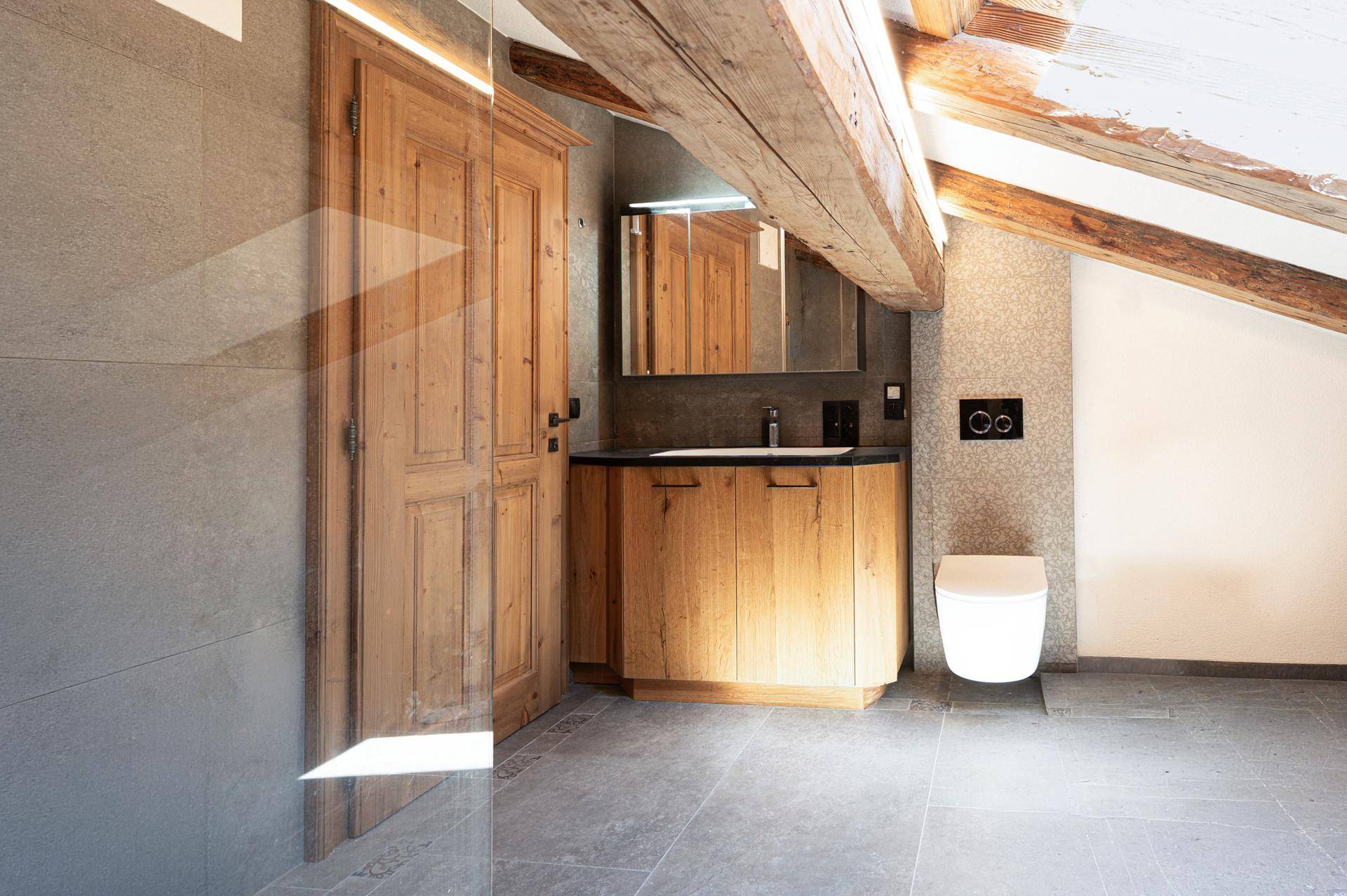 Modernes Badezimmer im Dachgeschoss mit Holztür, Waschtisch und Wand-WC unter Holzbalken.