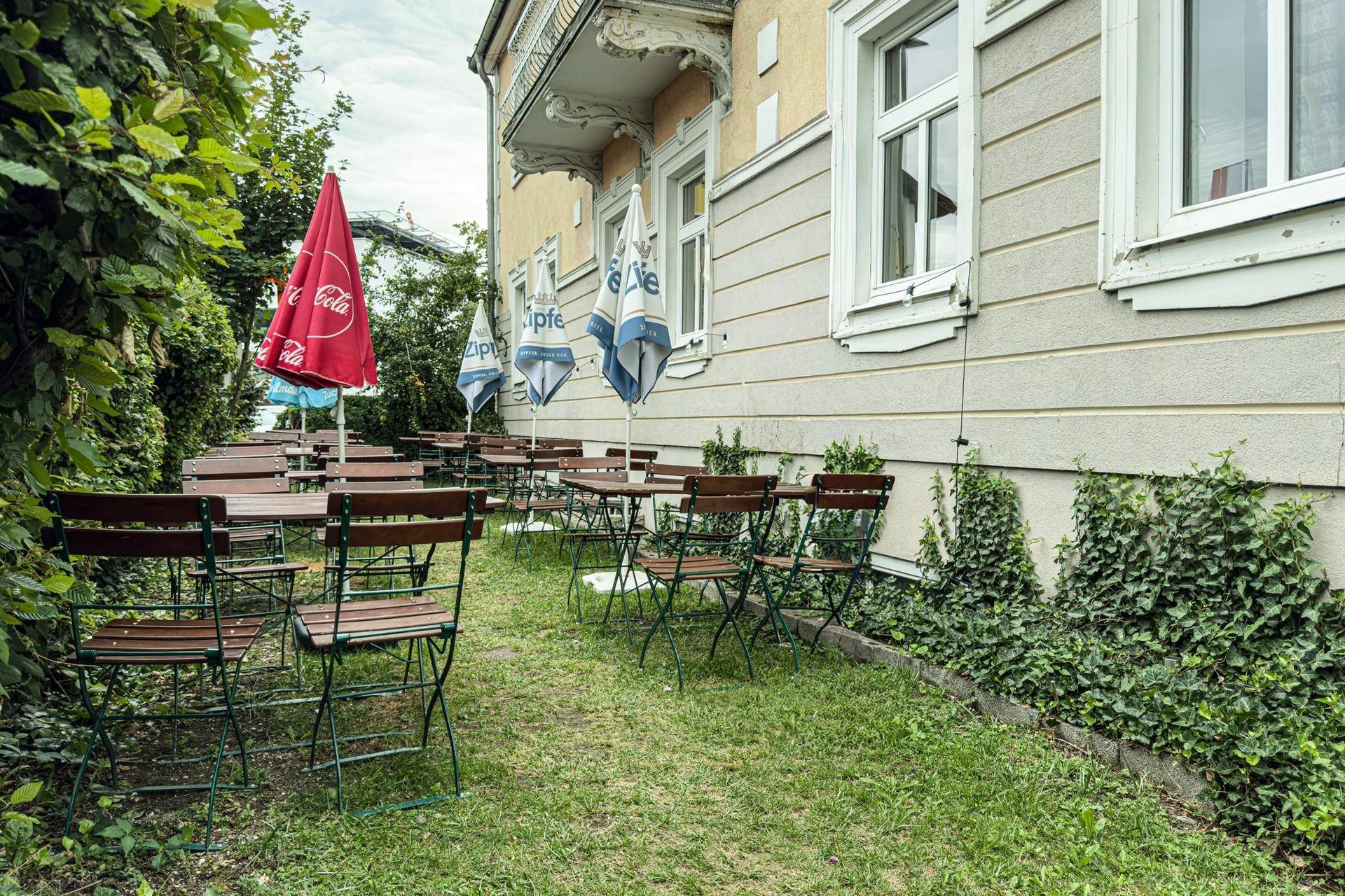 Außenbereich mit Tischen, Stühlen und Sonnenschirmen auf einer Rasenfläche neben dem Gebäude.