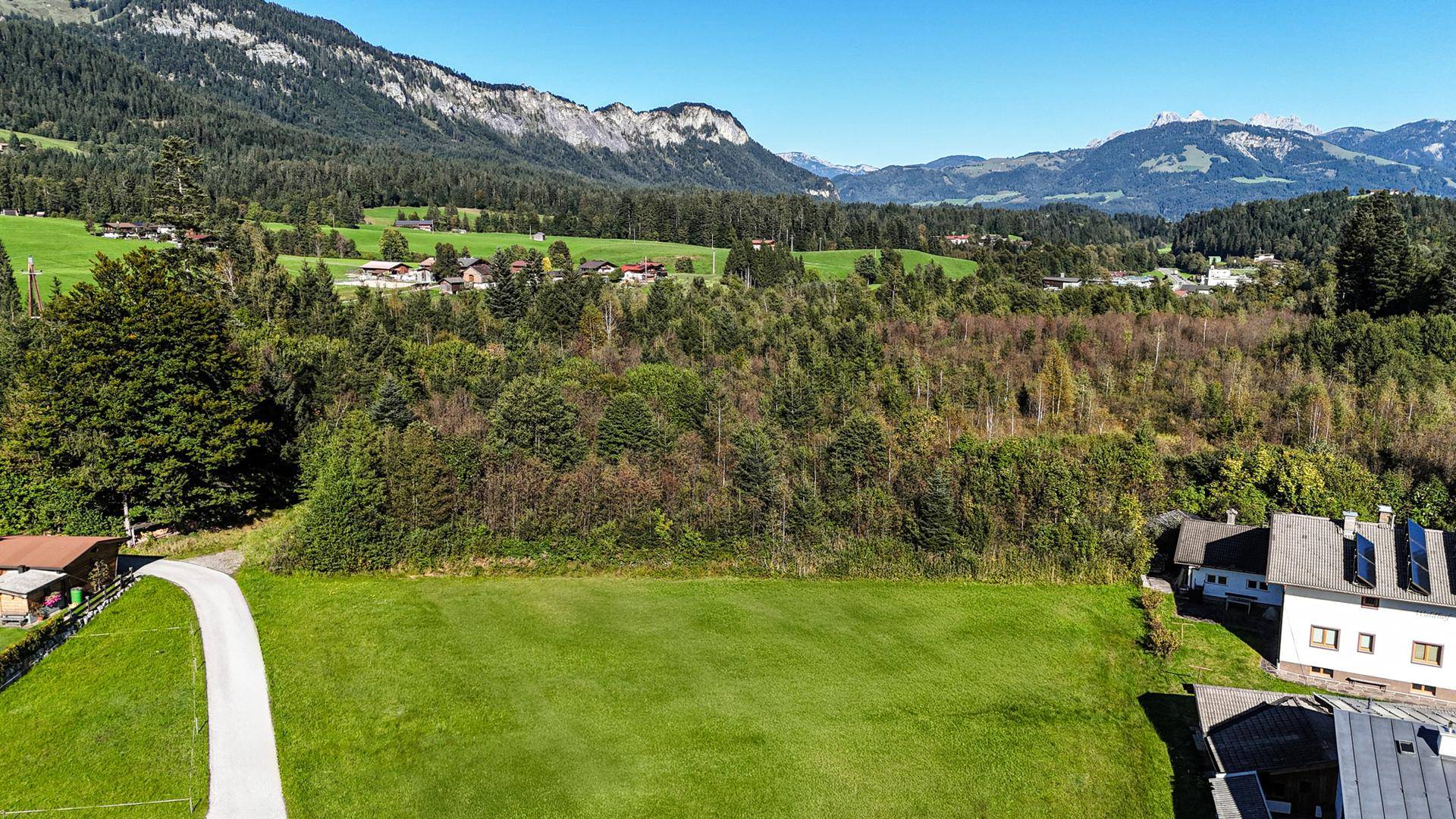 Luftaufnahme eines Grundstücks mit grüner Wiese, umgeben von Wald und Häusern, mit Bergen am Horizont.