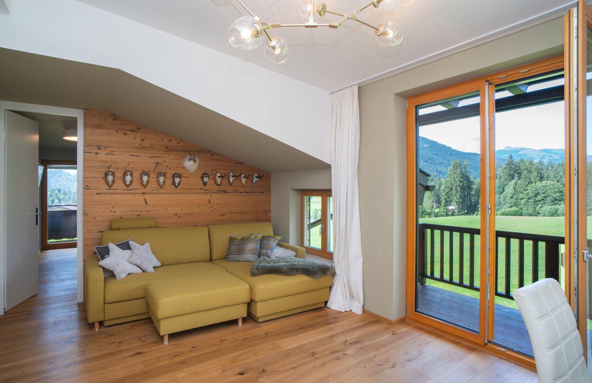Gemütliches Wohnzimmer mit Ecksofa, Holzwand und Zugang zu einem Balkon mit Bergblick.