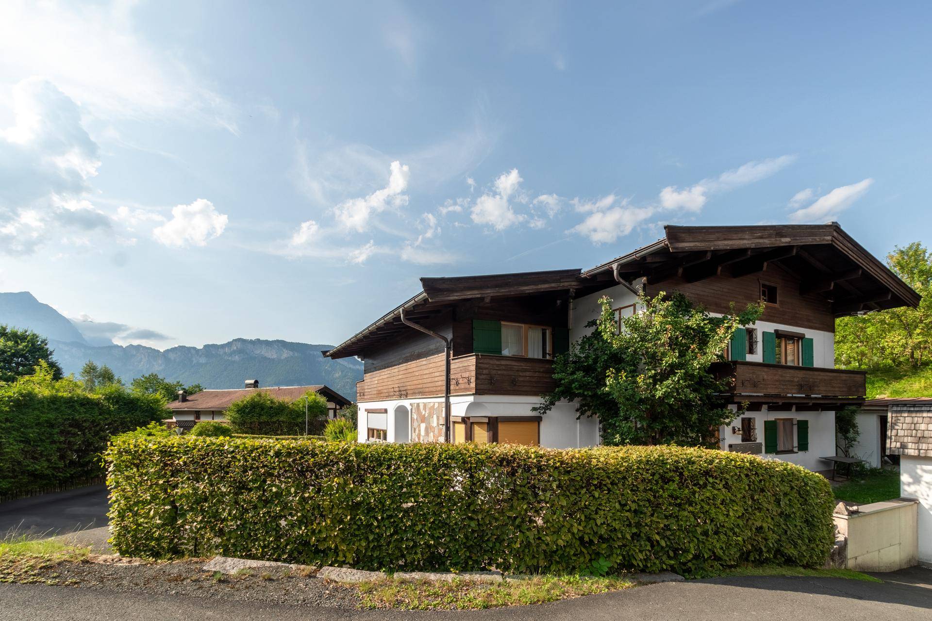 Alpenländisches Haus mit Holzelementen, grünen Fensterläden und Bergblick im Hintergrund.