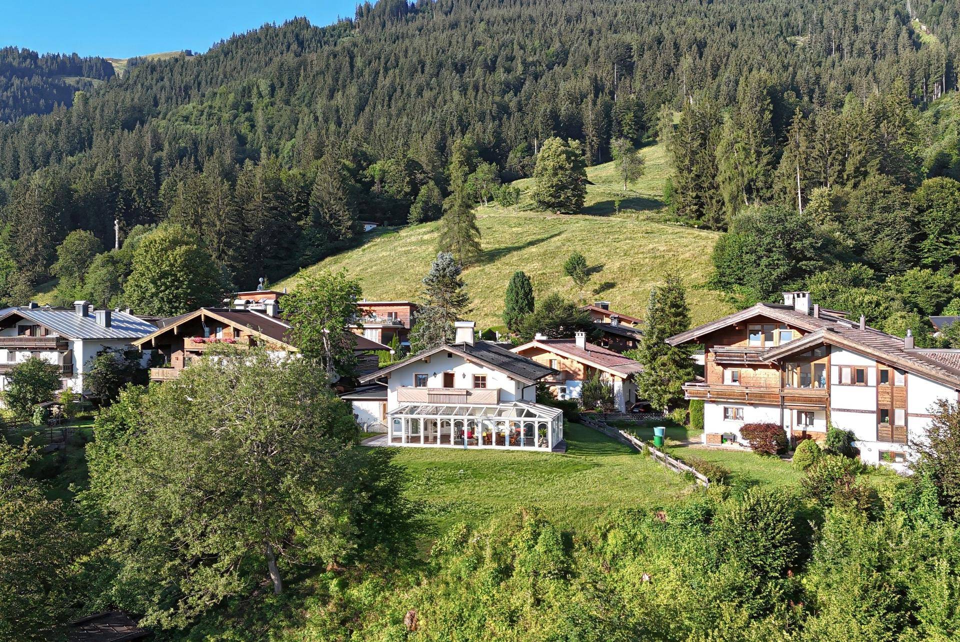 Mehrere Häuser in einer grünen, alpinen Landschaft mit bewaldeten Hügeln im Hintergrund.