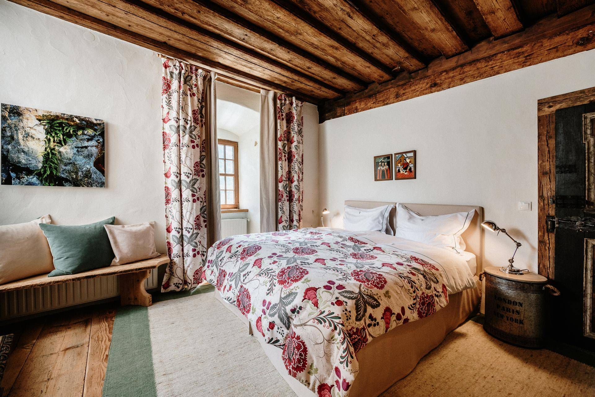 Helles Schlafzimmer mit Holzbalkendecke, floralen Vorhängen und gemütlichem Doppelbett.