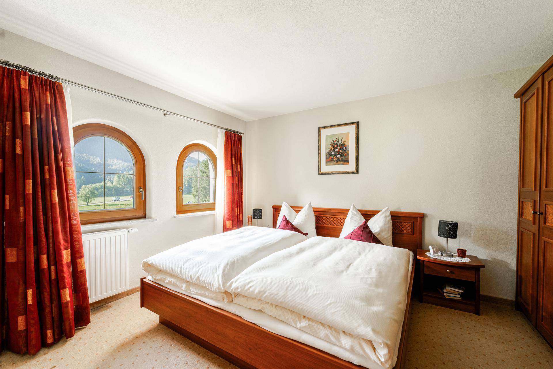Gemütliches Schlafzimmer mit Doppelbett, Holzmöbeln und Rundbogenfenstern mit Bergblick.
