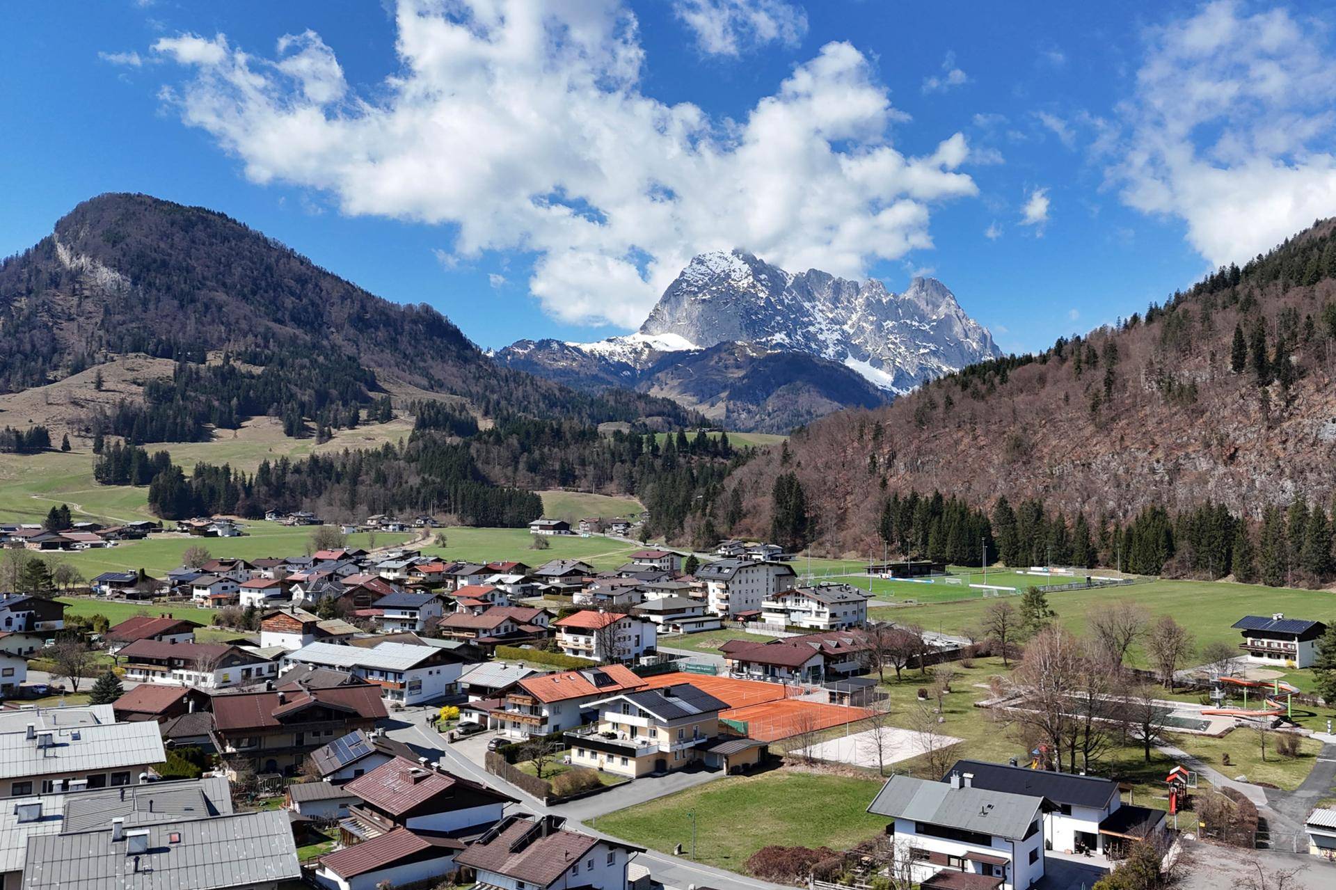 Panoramablick auf ein malerisches Dorf in den Alpen, umgeben von grünen Hügeln und hohen Bergen.