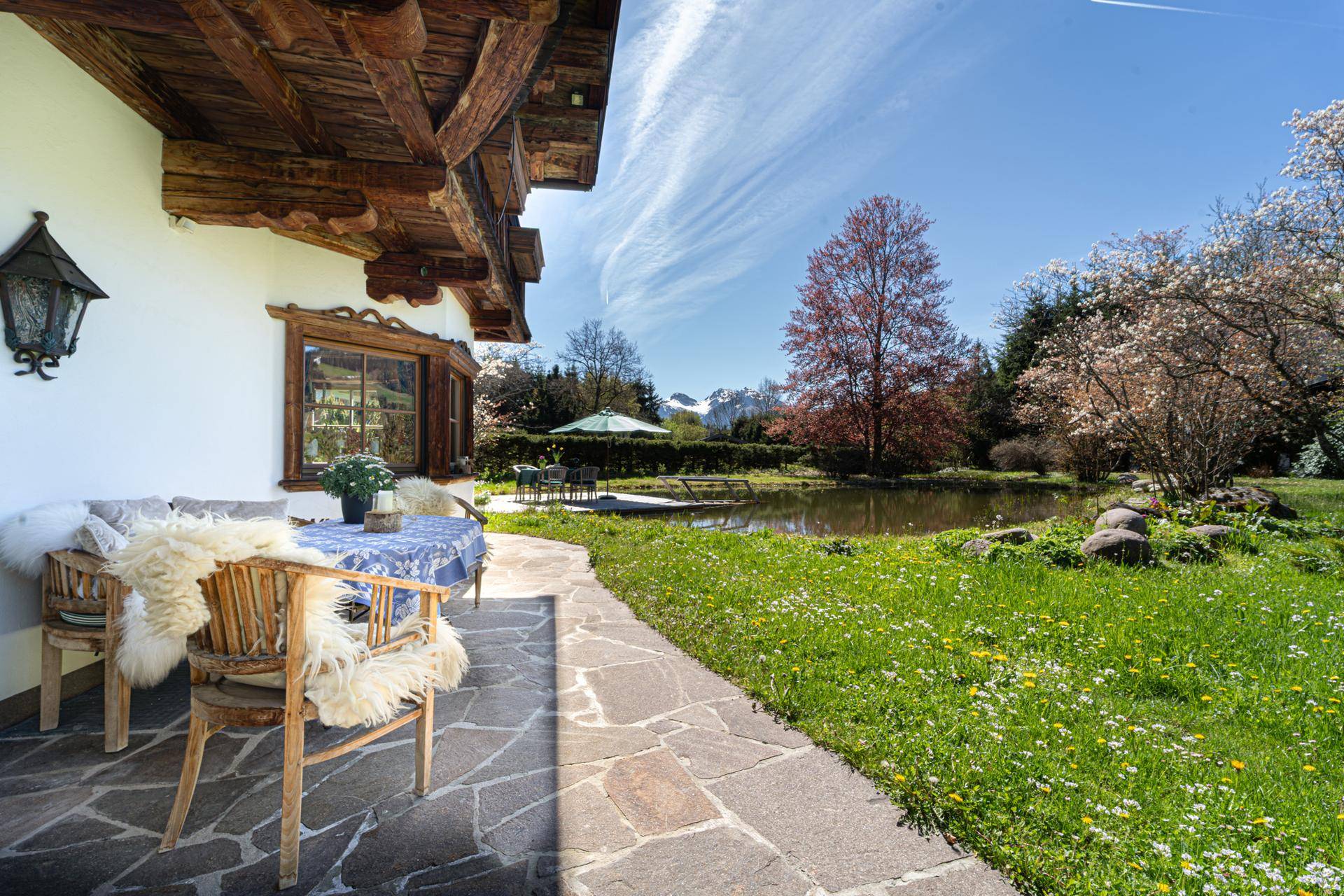 Sonniger Terrassenbereich mit Steinfliesen und Gartenmöbeln, umgeben von grüner Wiese und einem Teich.