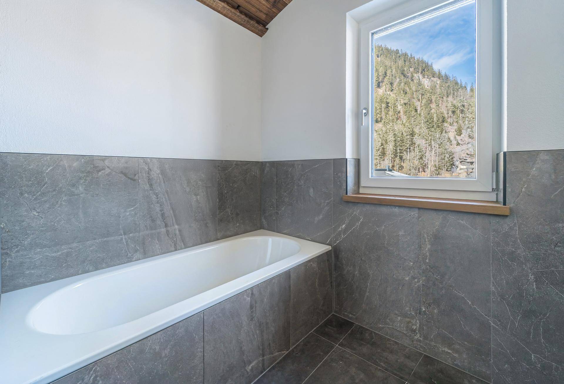 Modernes Badezimmer mit grauen Fliesen, Badewanne und Fenster mit Bergblick.