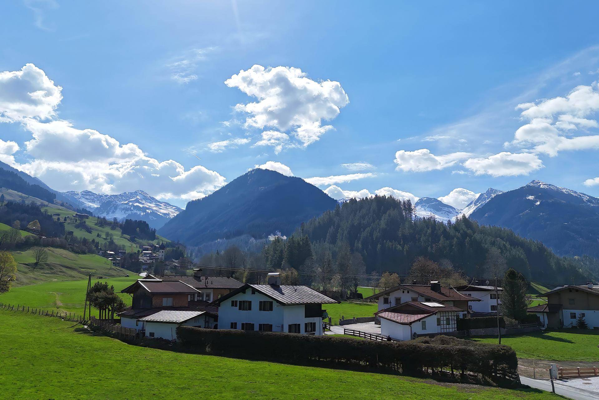 Panoramablick auf ein malerisches Dorf in den Alpen, umgeben von grünen Hügeln und schneebedeckten Bergen.