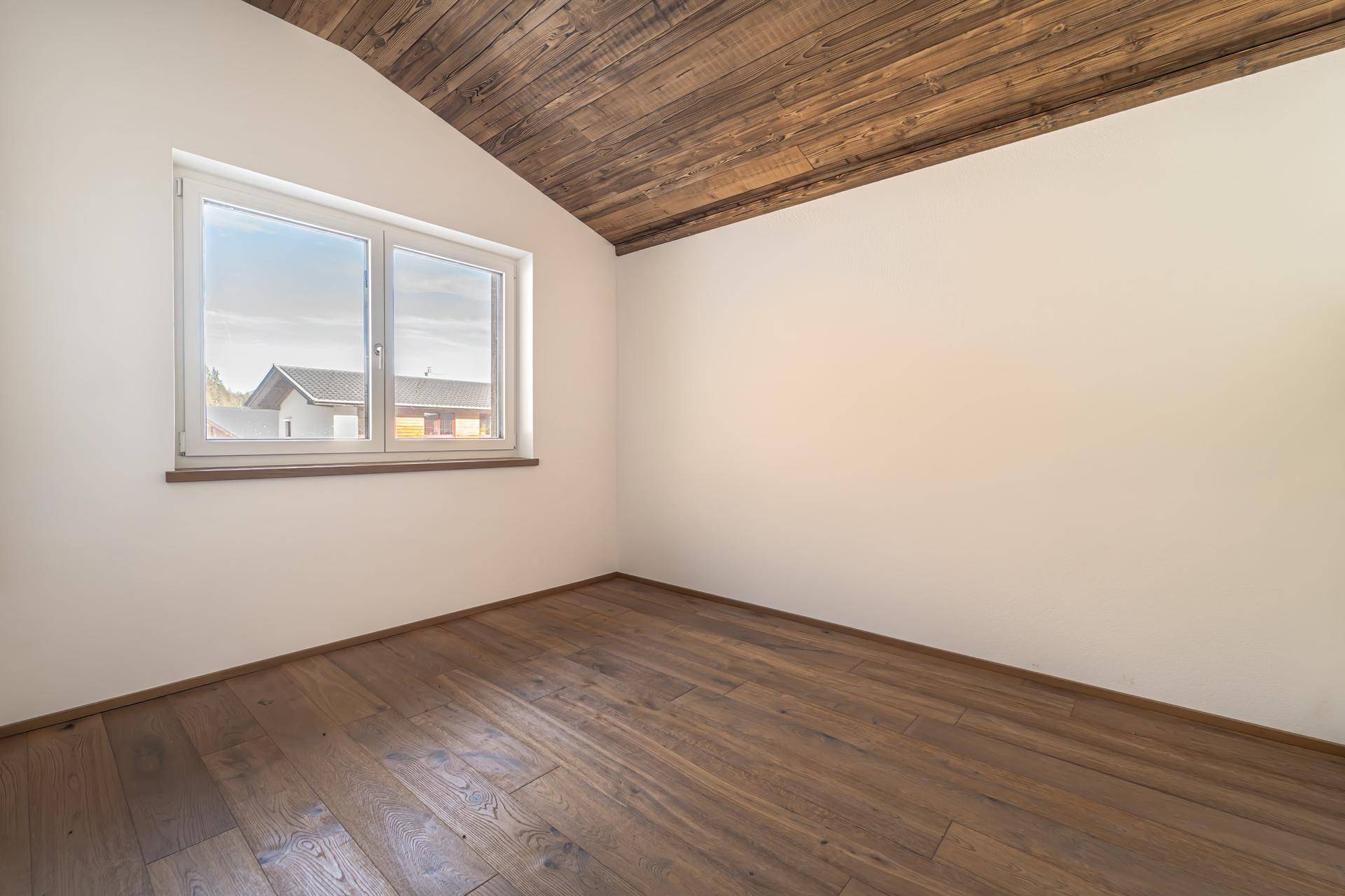 Heller Raum mit Holzboden, schräger Holzdecke und einem Fenster mit Blick auf die Umgebung.