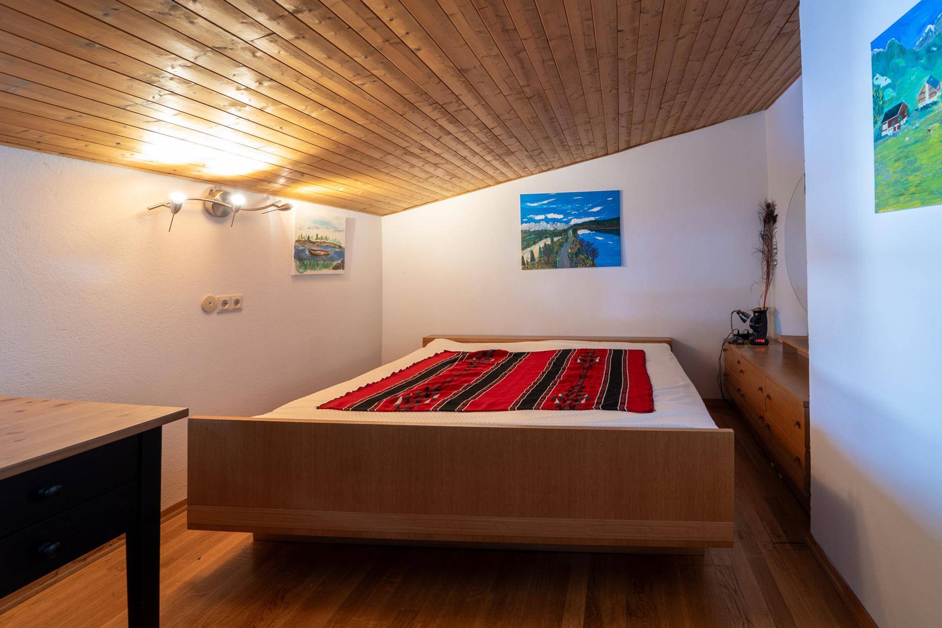 Schlafzimmer mit Holzbalkendecke, Holzboden und farbenfrohen Bildern an der Wand.