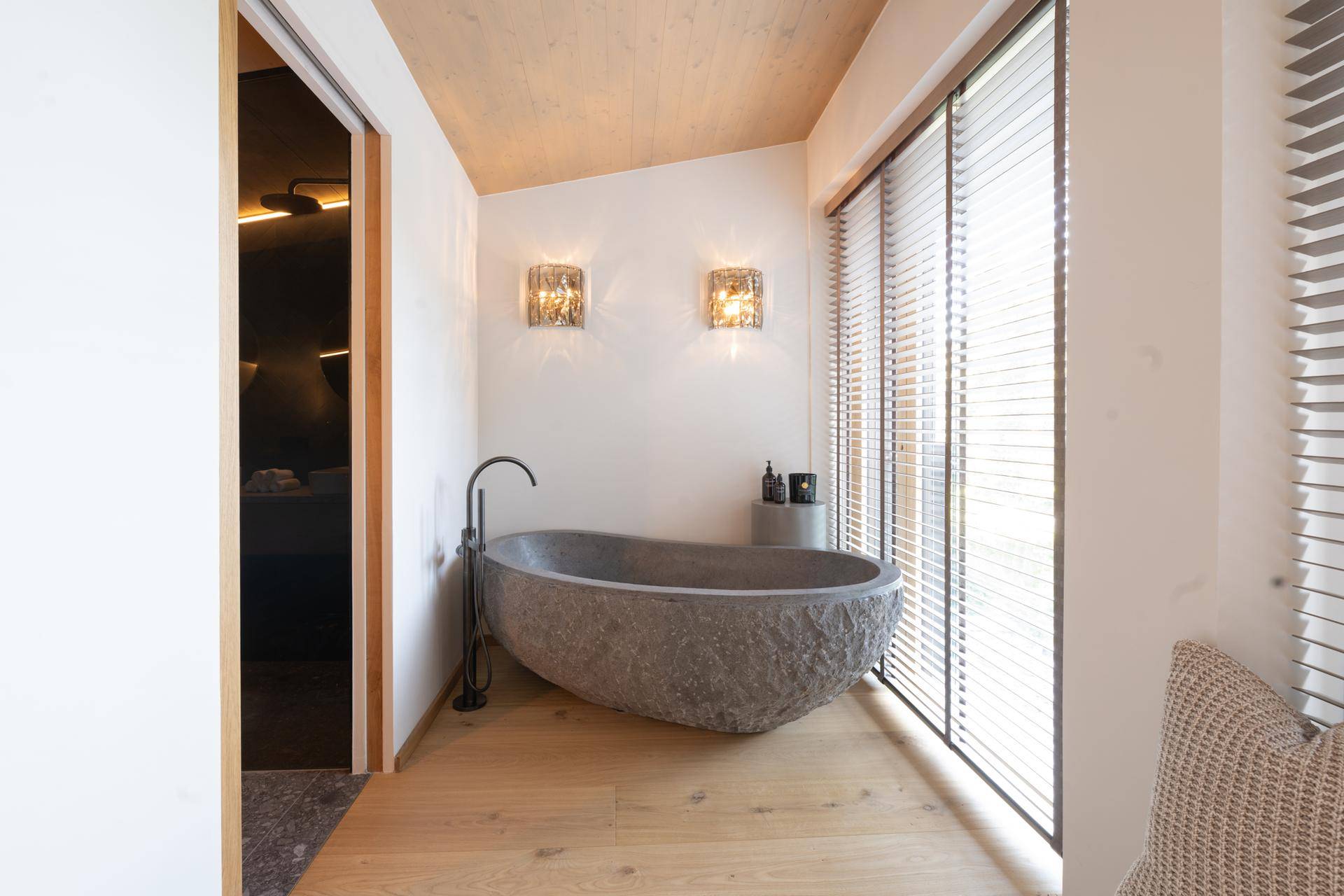 Elegantes Badezimmer mit freistehender Steinbadewanne, Holzboden und großen Fenstern.