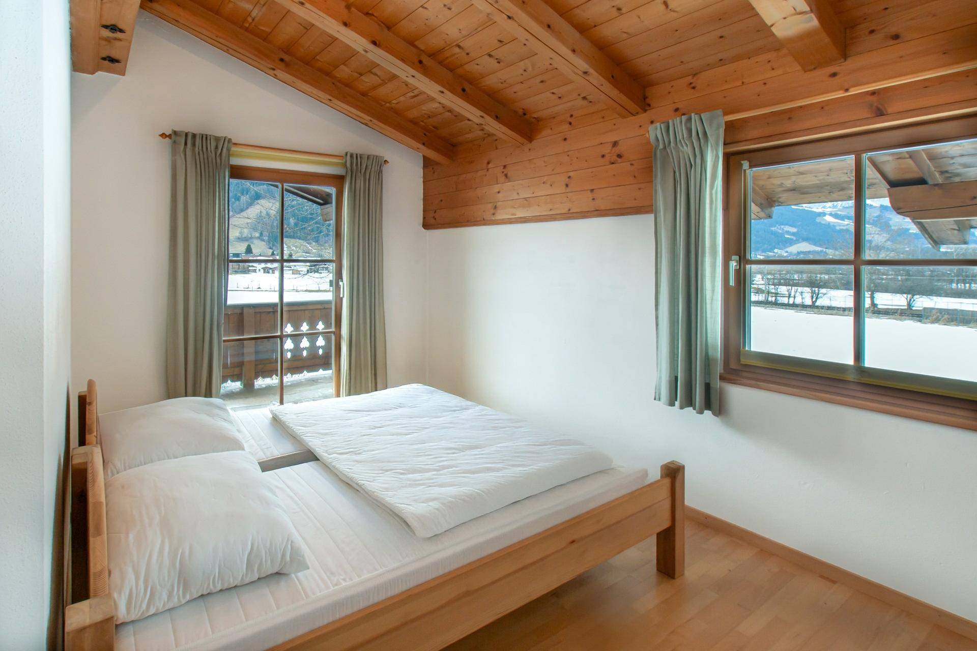Helles Schlafzimmer mit Holzbett, schrägen Decken und Blick auf die verschneite Berglandschaft.