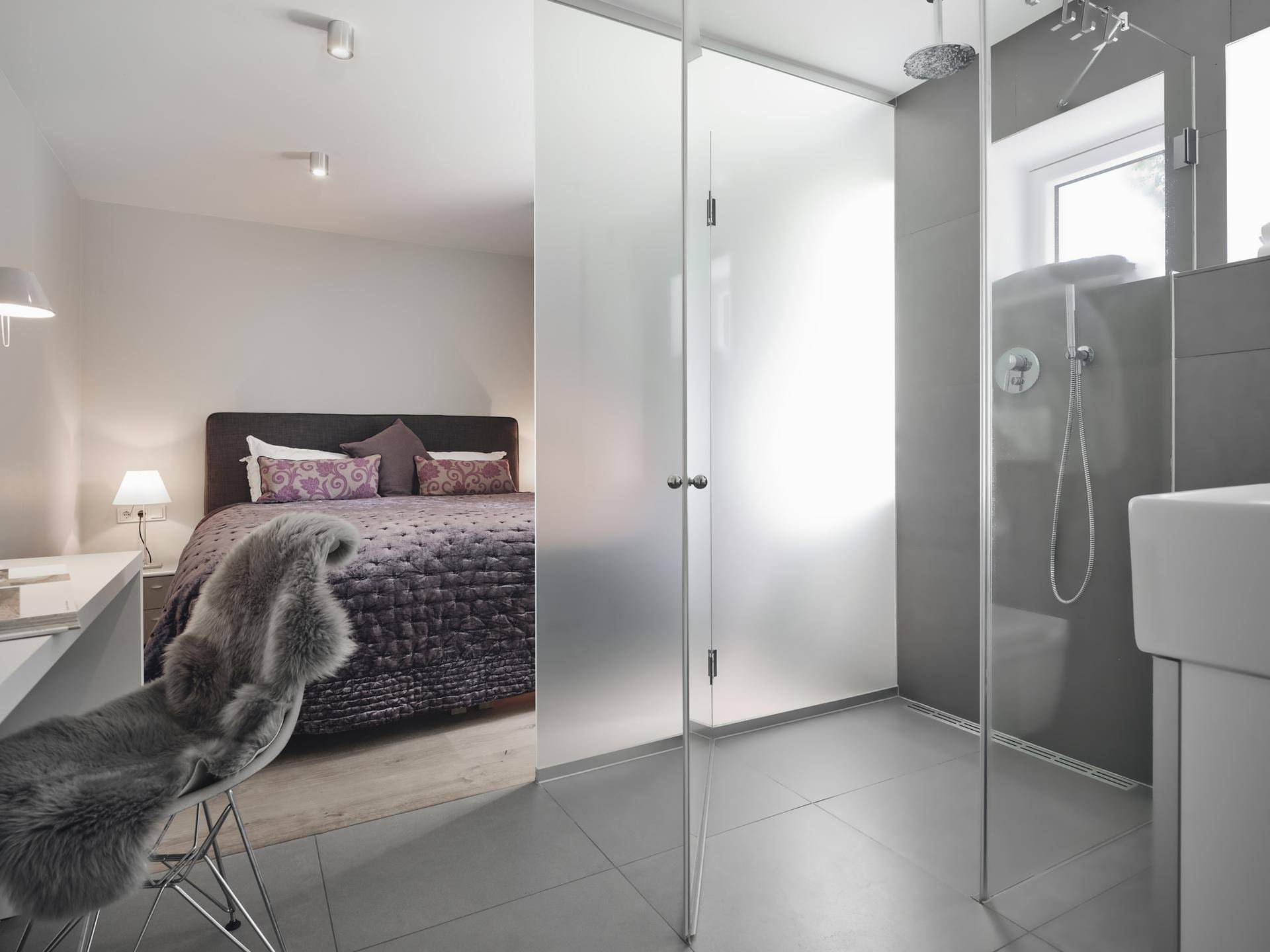 Schlafzimmer mit offenem Badbereich, ausgestattet mit einer modernen begehbaren Dusche und Fliesenboden.