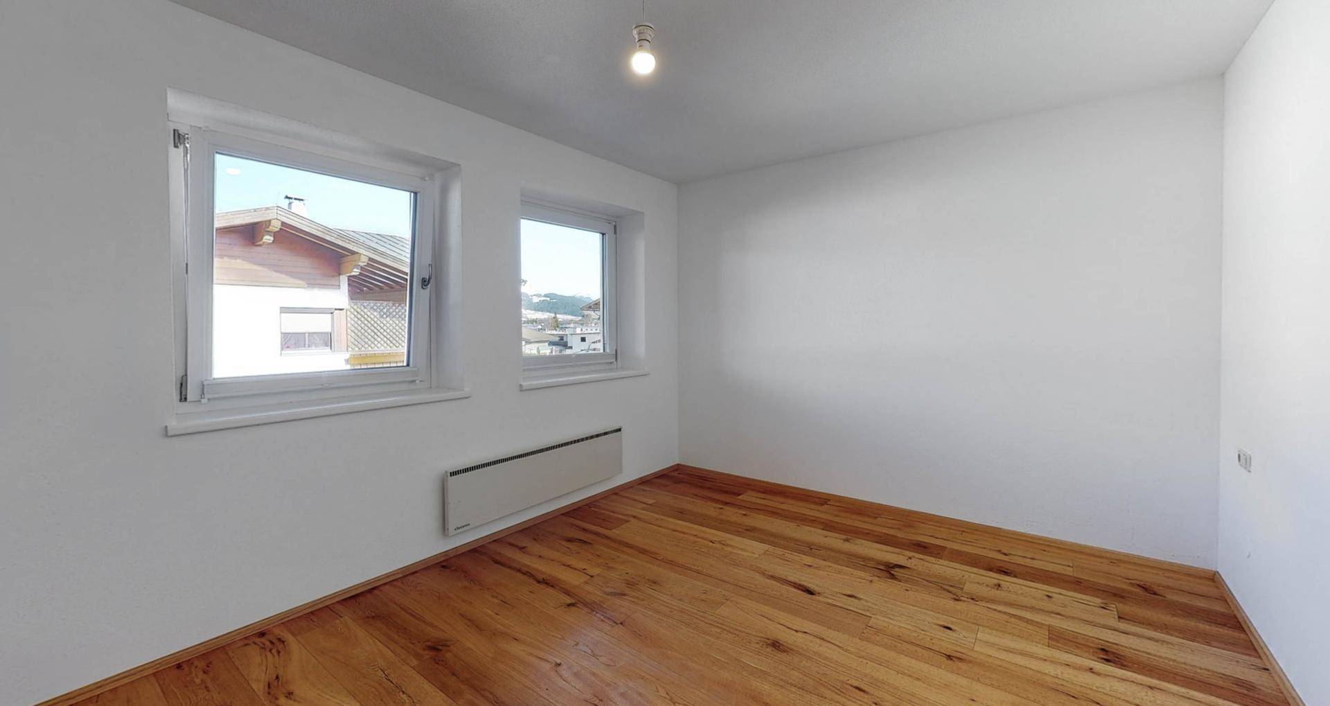 Leeres Zimmer mit Holzboden, weißen Wänden und zwei Fenstern.