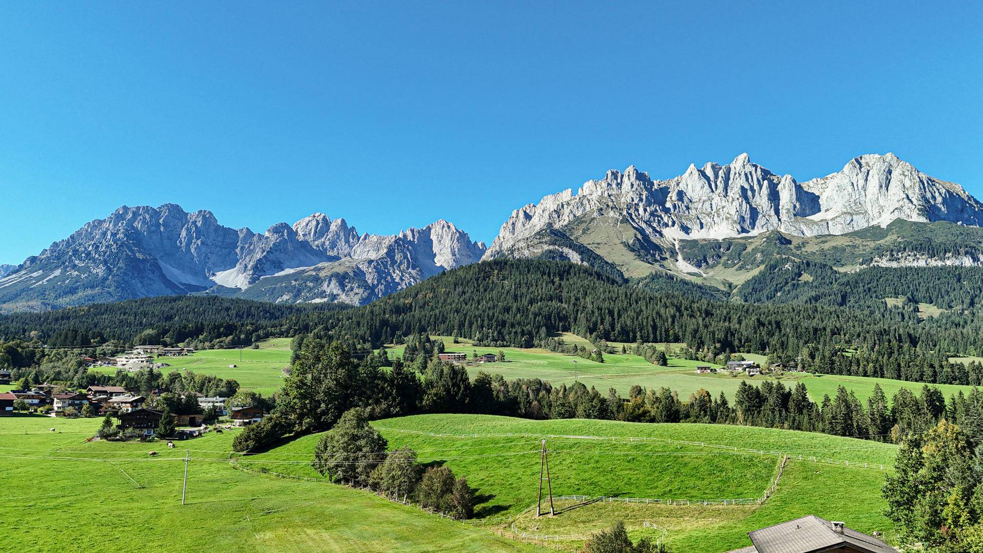 Idyllische Landschaft mit sanften grünen Hügeln, verstreuten Häusern und einer majestätischen Bergkette unter klarem Himmel.