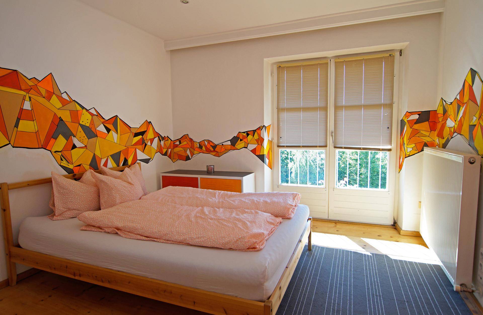 Schlafzimmer mit Doppelbett, Holzboden und farbenfroher Wandgestaltung in Orange- und Gelbtönen.