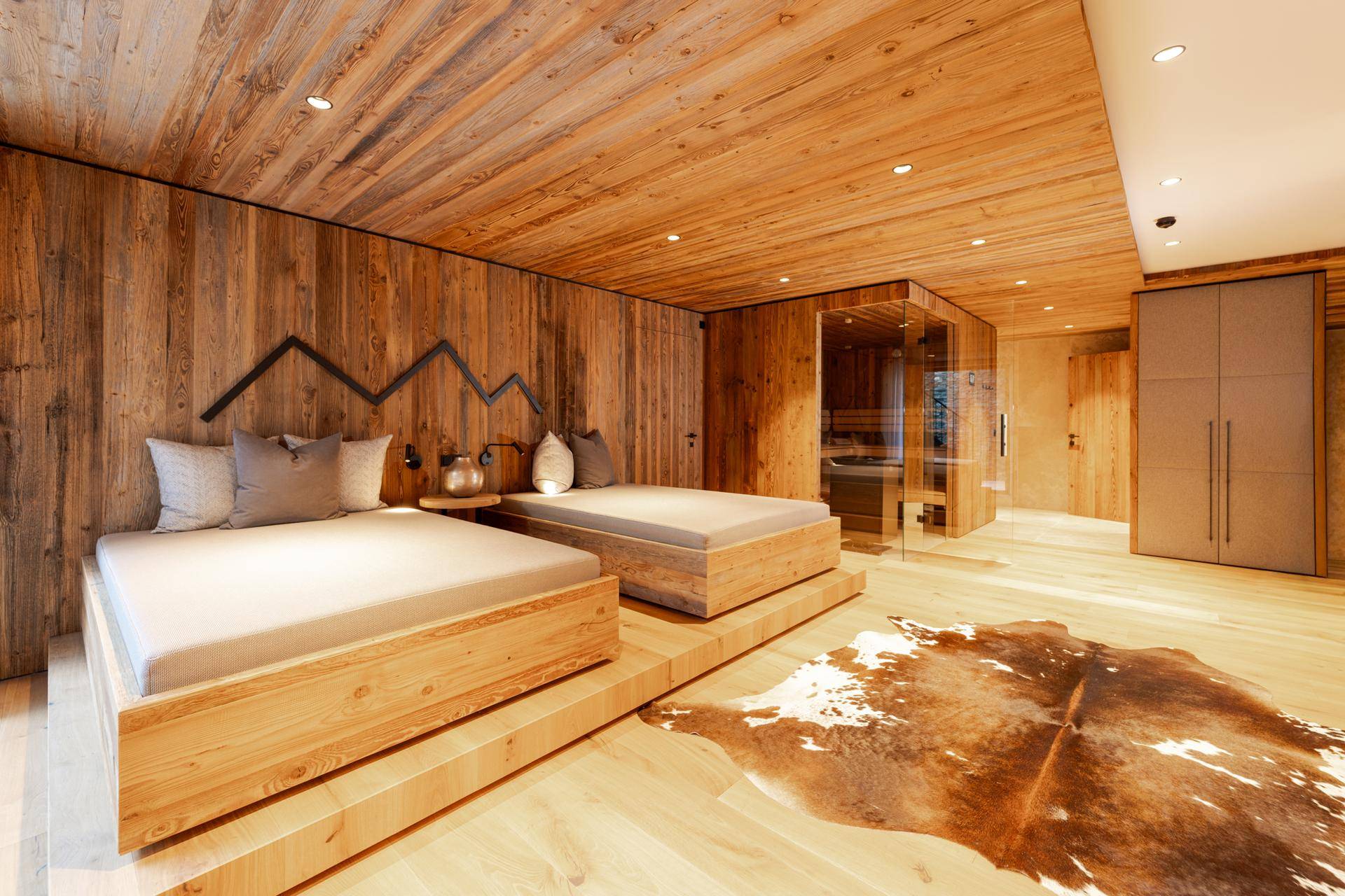 Rustikales Schlafzimmer mit zwei Einzelbetten auf Holzpodesten, Kuhfellteppich und integrierter Sauna.
