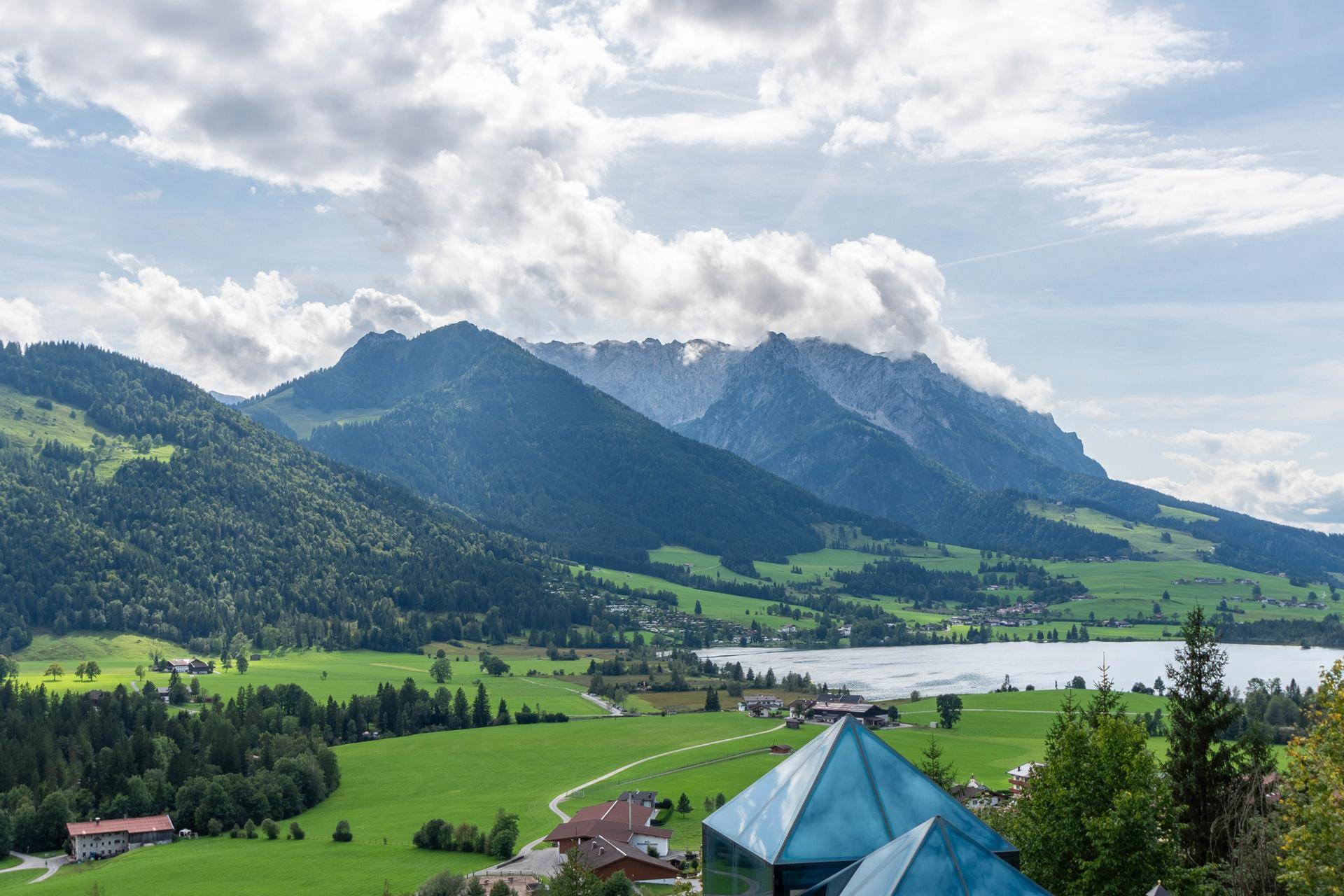 Atemberaubende Landschaft mit majestätischen Bergen, einem ruhigen See und einem Dorf, im Vordergrund eine moderne Glasstruktur.
