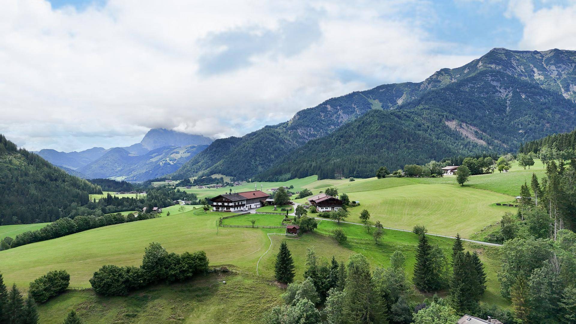 Weitläufige grüne Hügellandschaft mit vereinzelten Häusern und majestätischen Bergen im Hintergrund.