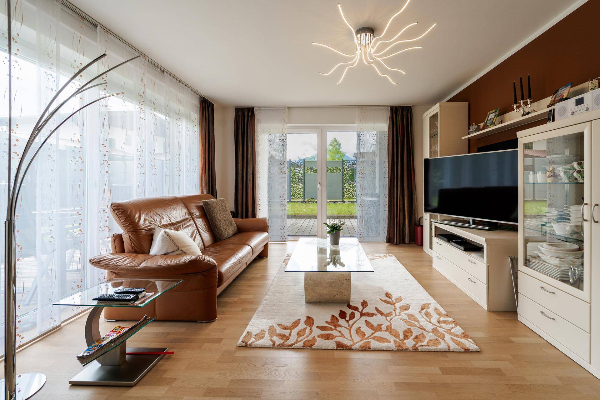 Helles Wohnzimmer mit braunem Ledersofa, modernem Teppich und Zugang zur Terrasse.