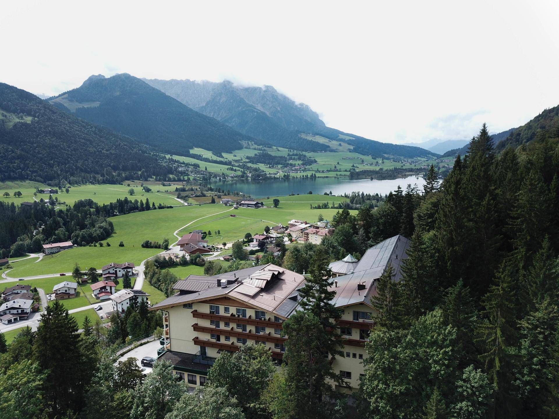 Panoramablick auf ein Gebäudeensemble mit See und majestätischen Bergen im Hintergrund, umgeben von grünen Wiesen.