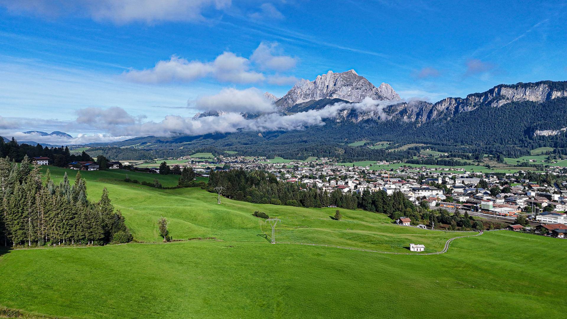 Panoramablick auf die weite grüne Landschaft mit Wäldern, einem Dorf und imposanten Bergen unter blauem Himmel.