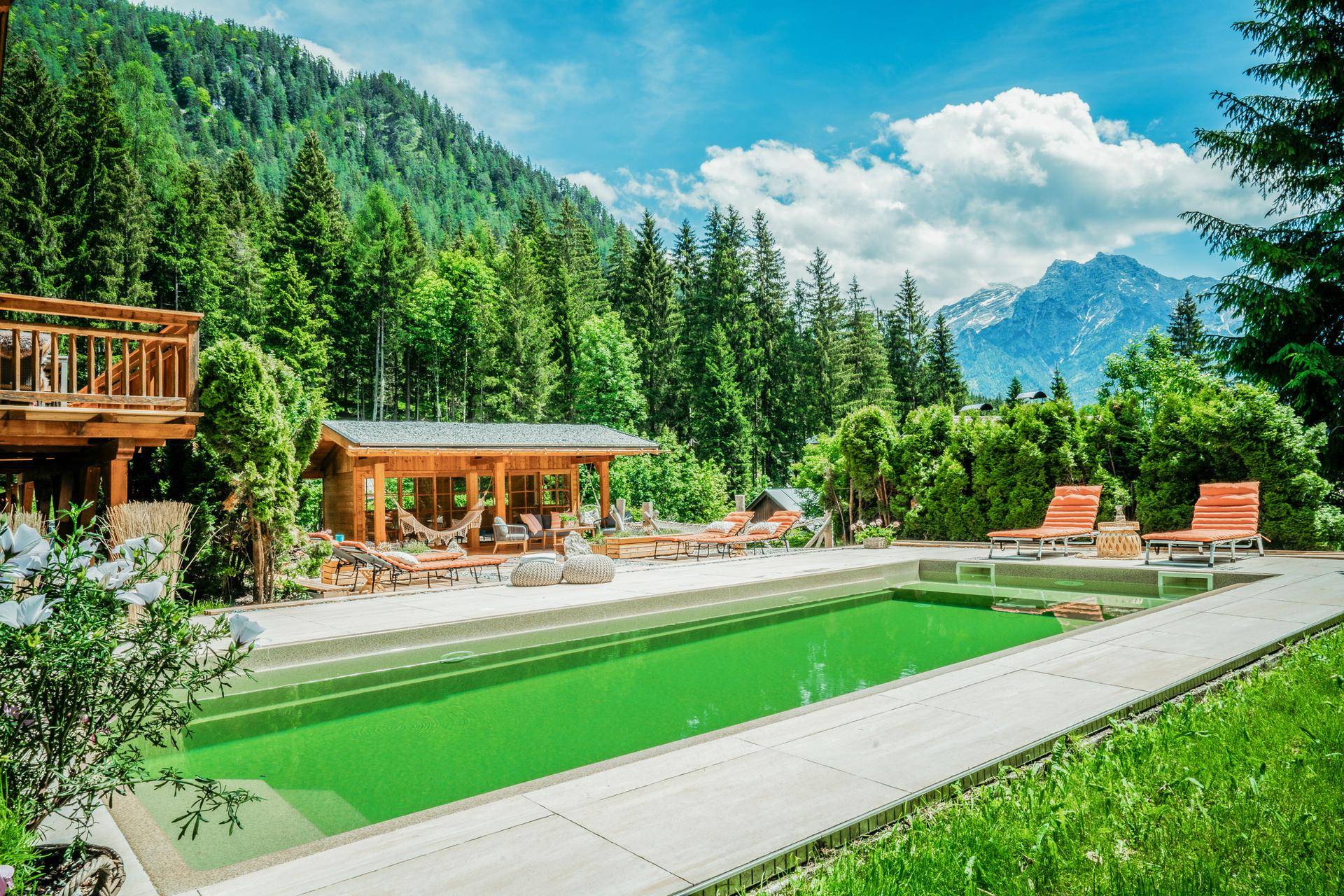 Großer Außenpool in einer grünen Berglandschaft mit Liegestühlen und einem Gartenhaus.